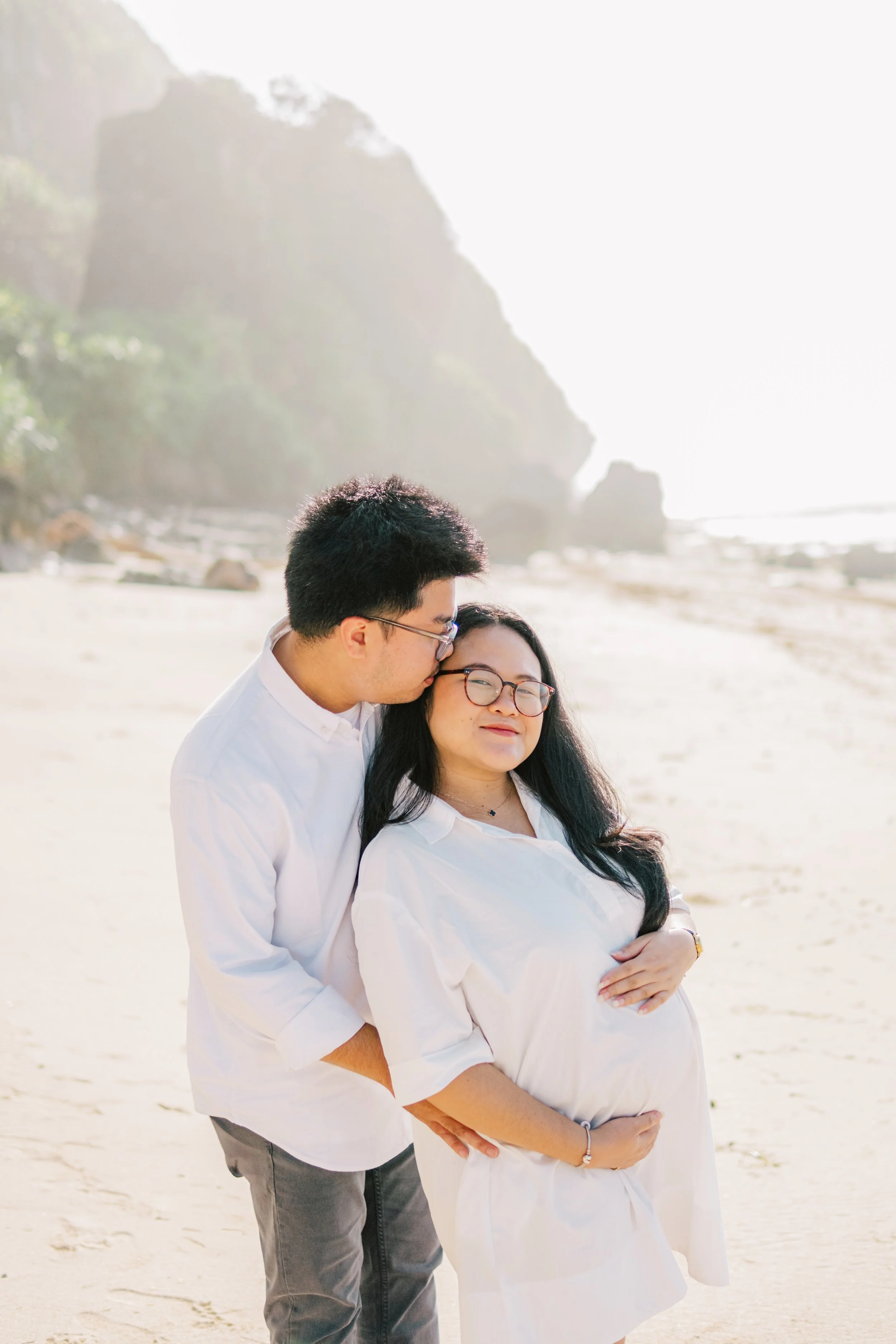 A man and woman, both dressed in white, standing on a beach. The man is kissing the woman on the forehead while she holds her pregnant belly and smiles gently. The background features large rocky formations and the sky is bright.