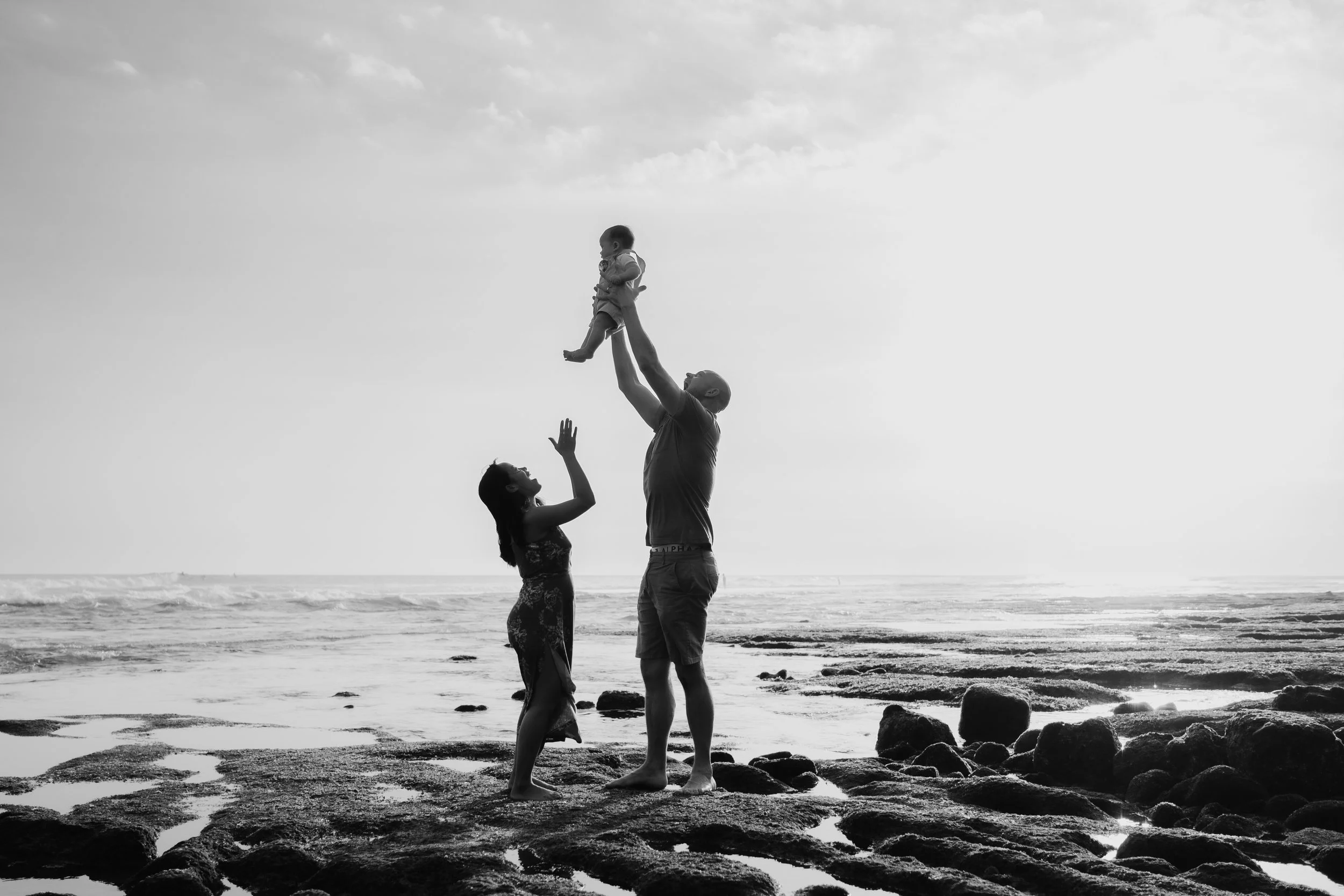 A black-and-white photo of a family at the beach, with a father holding a child high in the air, a woman reaching up towards the child, and rocks on the shoreline in the background.