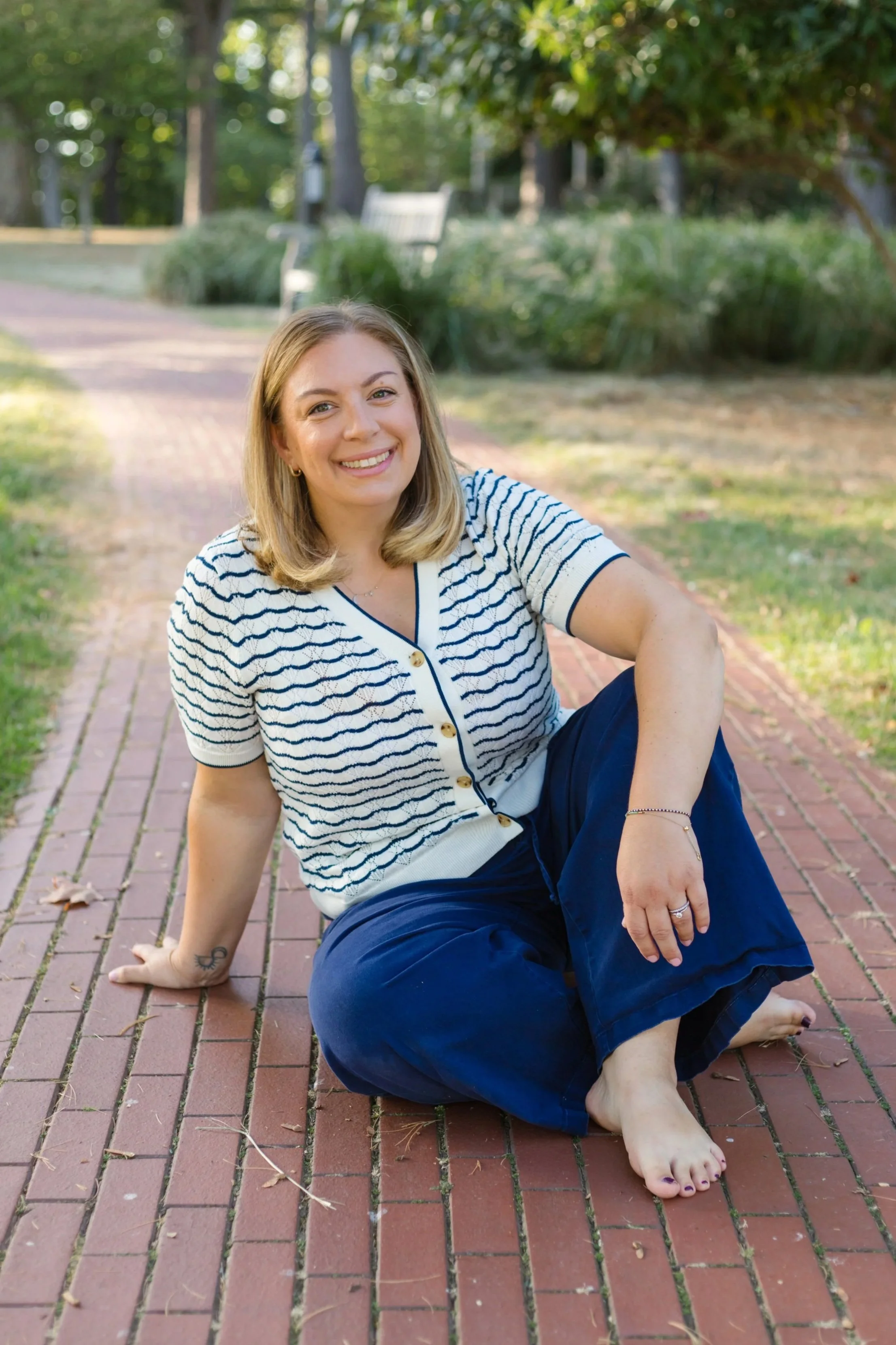 A woman with long blonde hair and glasses stands indoors, wearing a black shirt and a long necklace. She is leaning against a white lattice railing.