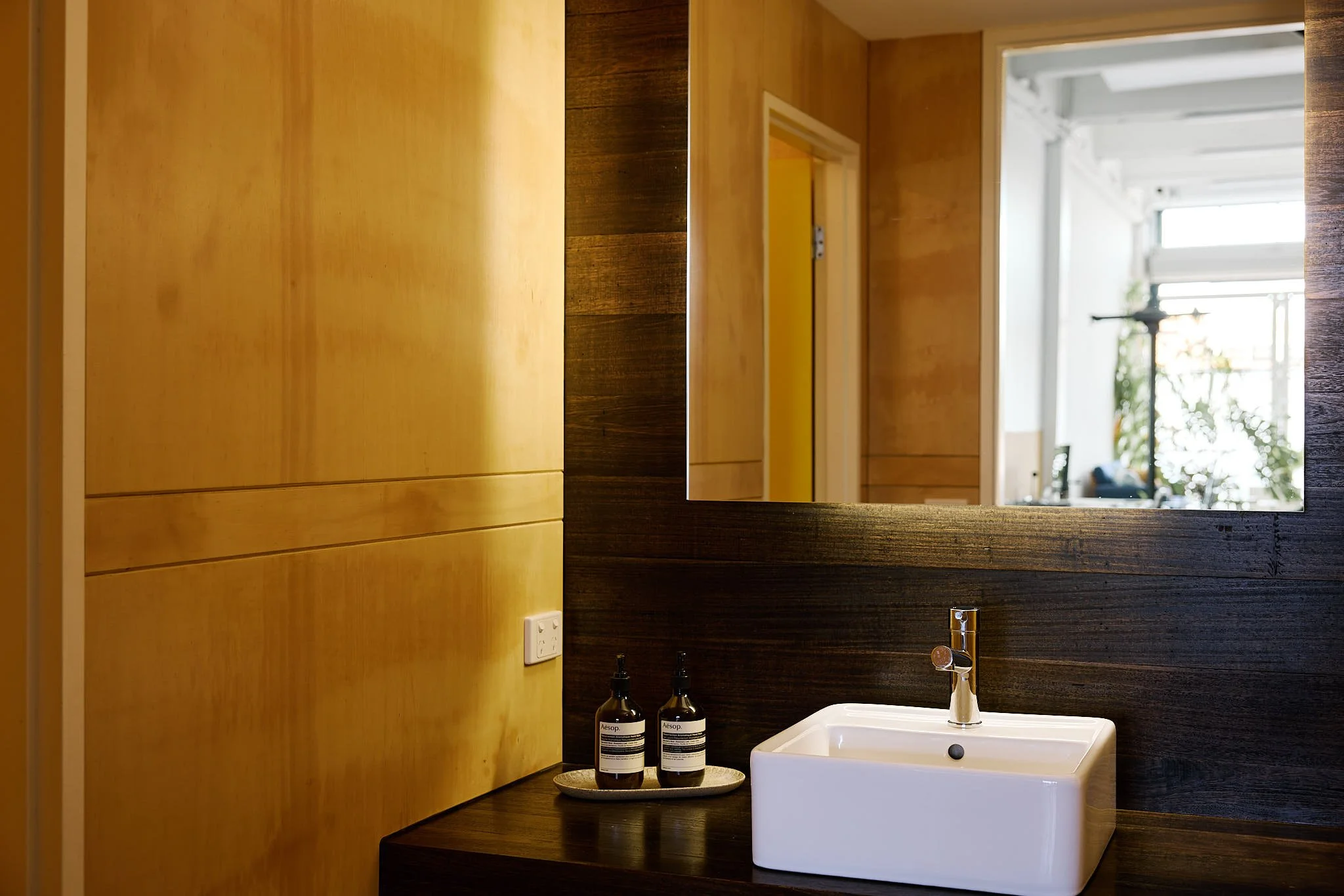 Modern bathroom interior with wooden paneling, a large mirror, a white square sink, and bottles of soap or lotion on the counter.