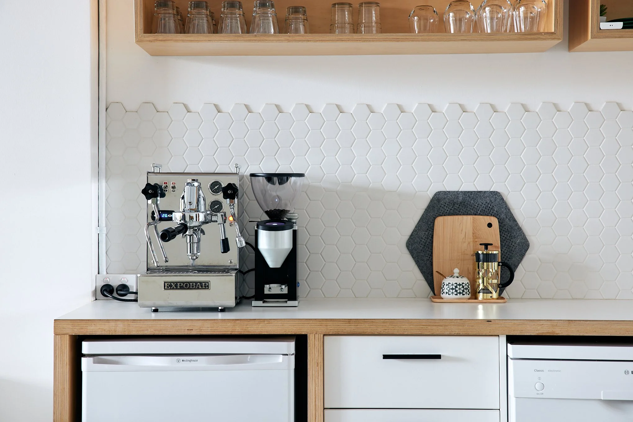 Modern kitchen with espresso machine, coffee grinder, and French press on a white countertop against hexagonal tiled backsplash.