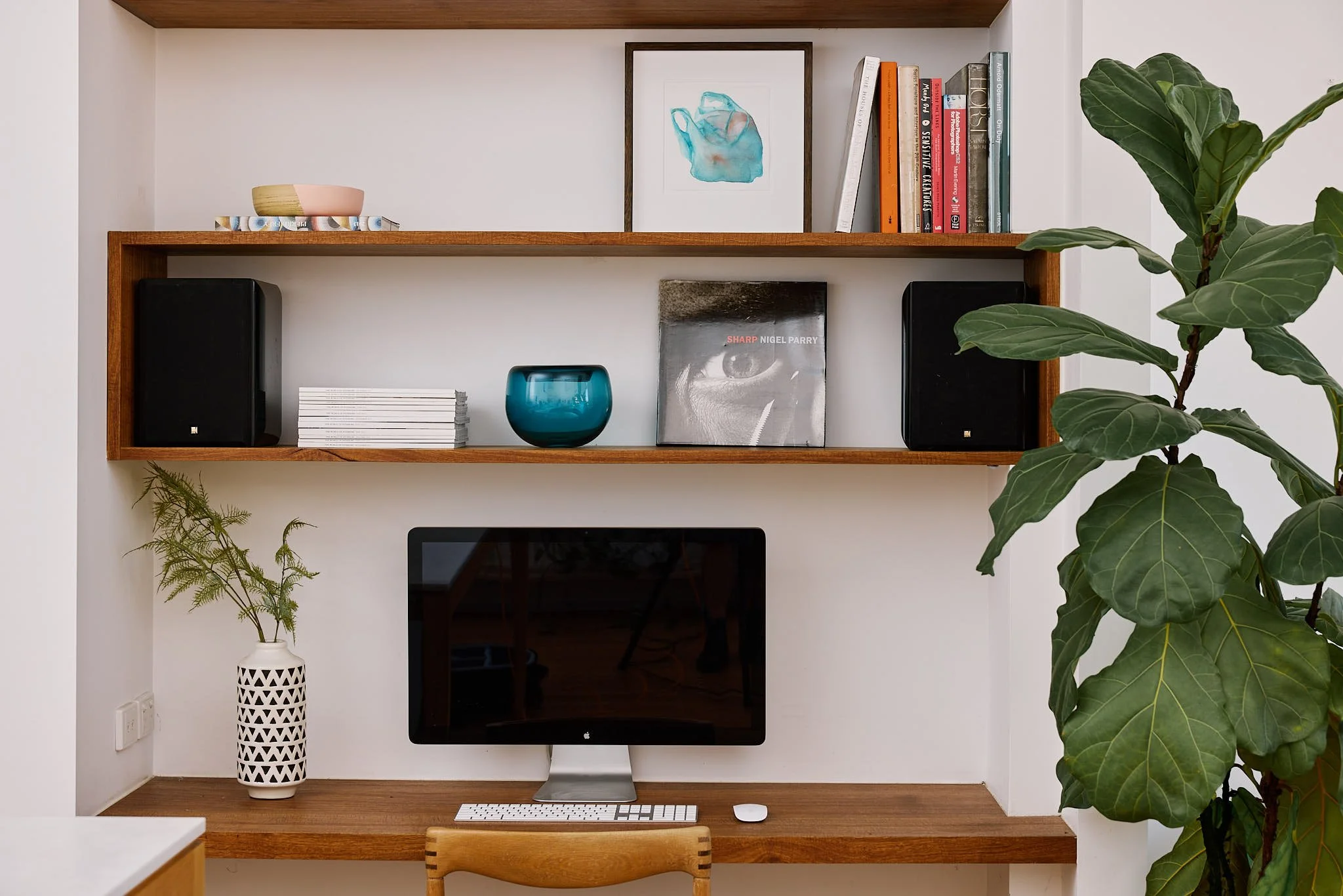 Home office setup with wooden shelves, a computer monitor, keyboard, mouse, books, speakers, a framed picture, decorative bowl, plant, and magazines.