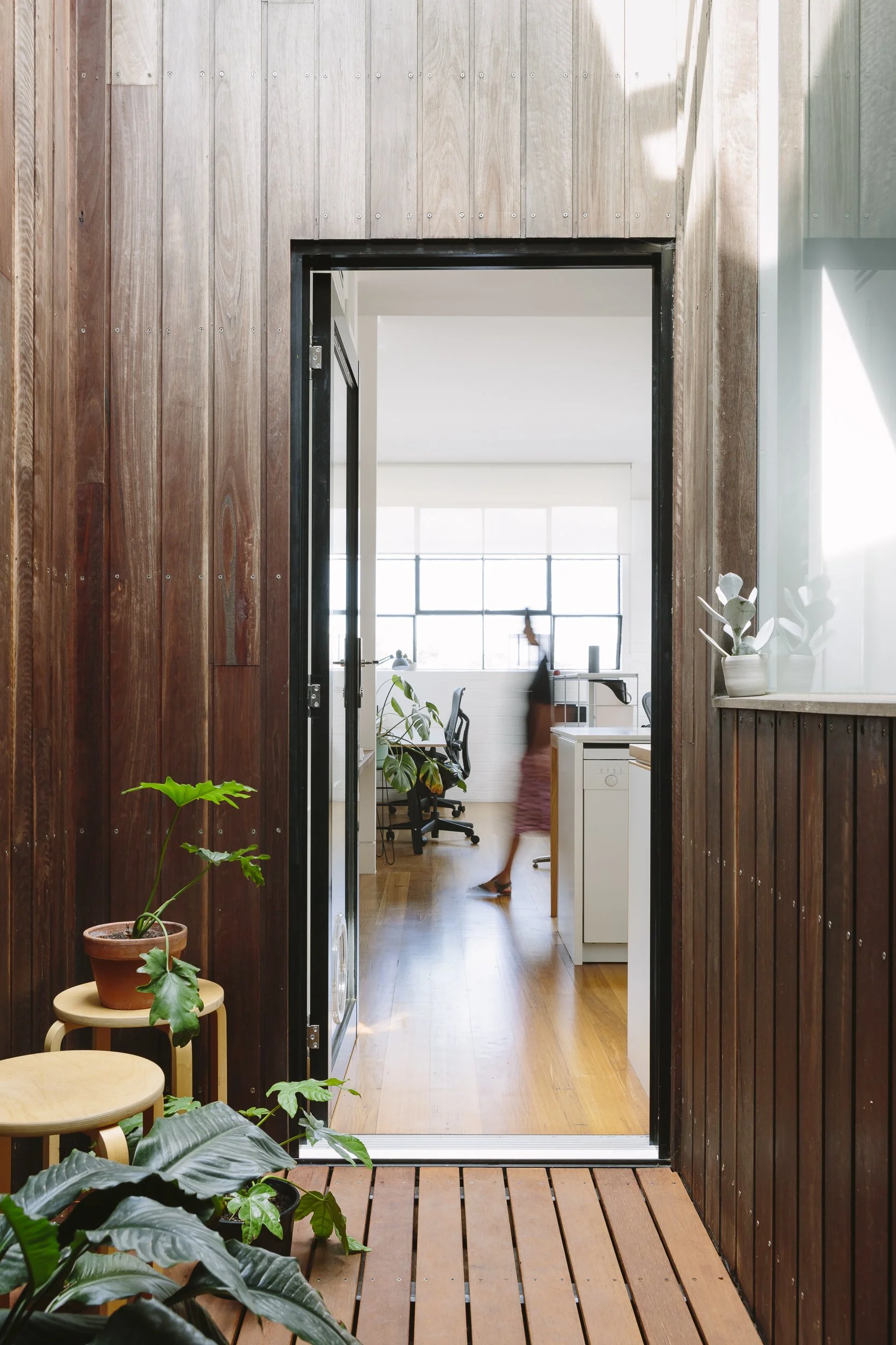 Open wooden door leading to a bright interior space with potted plants and a blurred person walking inside.