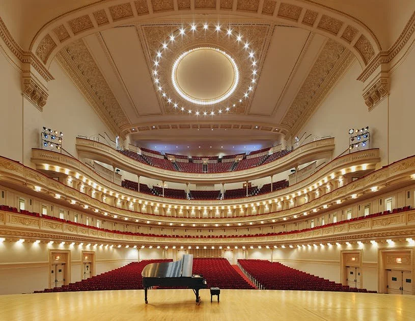 Empty concert hall with a grand piano on stage, ornate balconies, red theater seats, and a large decorative ceiling with a circular chandelier.