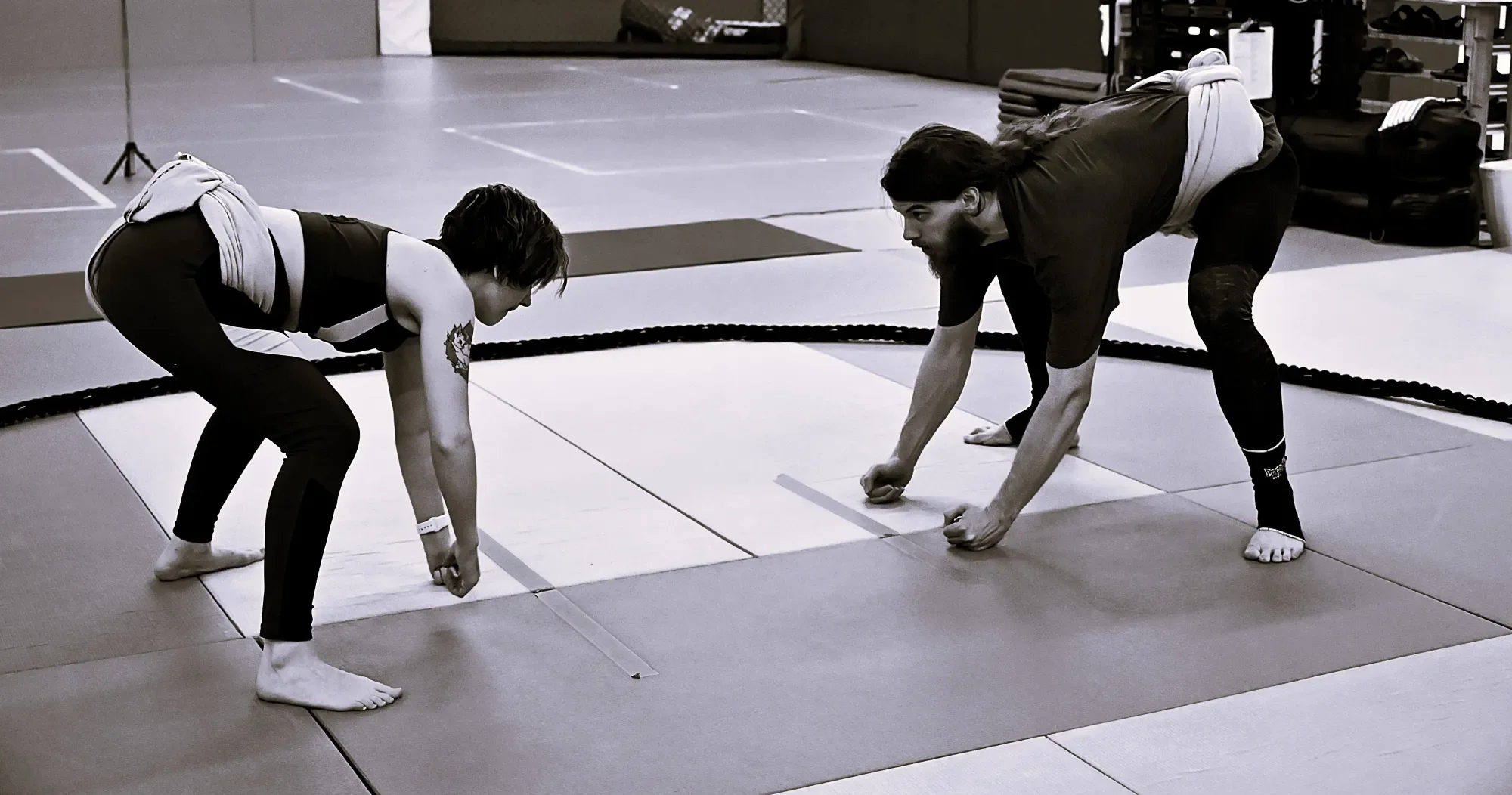 A woman and a girl practicing wrestling on a mats in a gym, both in a crouched position facing each other, preparing to engage.