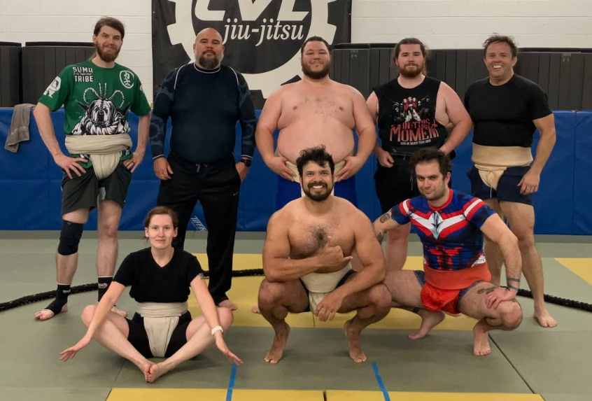Group of eight people in martial arts uniforms and casual clothing posing on a gym mat, with a jiu-jitsu themed banner in the background.