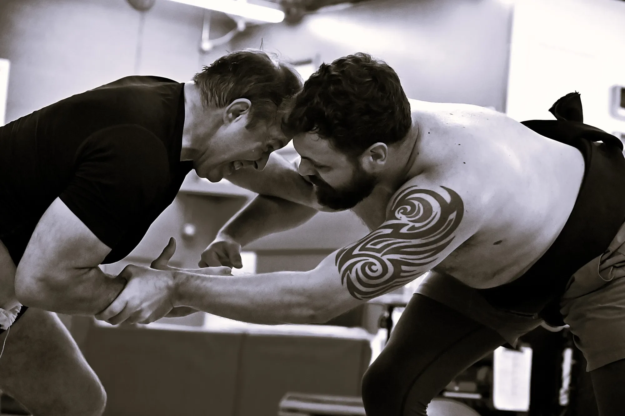 Two men are engaged in a wrestling match, one with a tribal tattoo on his upper arm, with their foreheads pressed together and hands gripping each other's arms, in a training or gym environment.