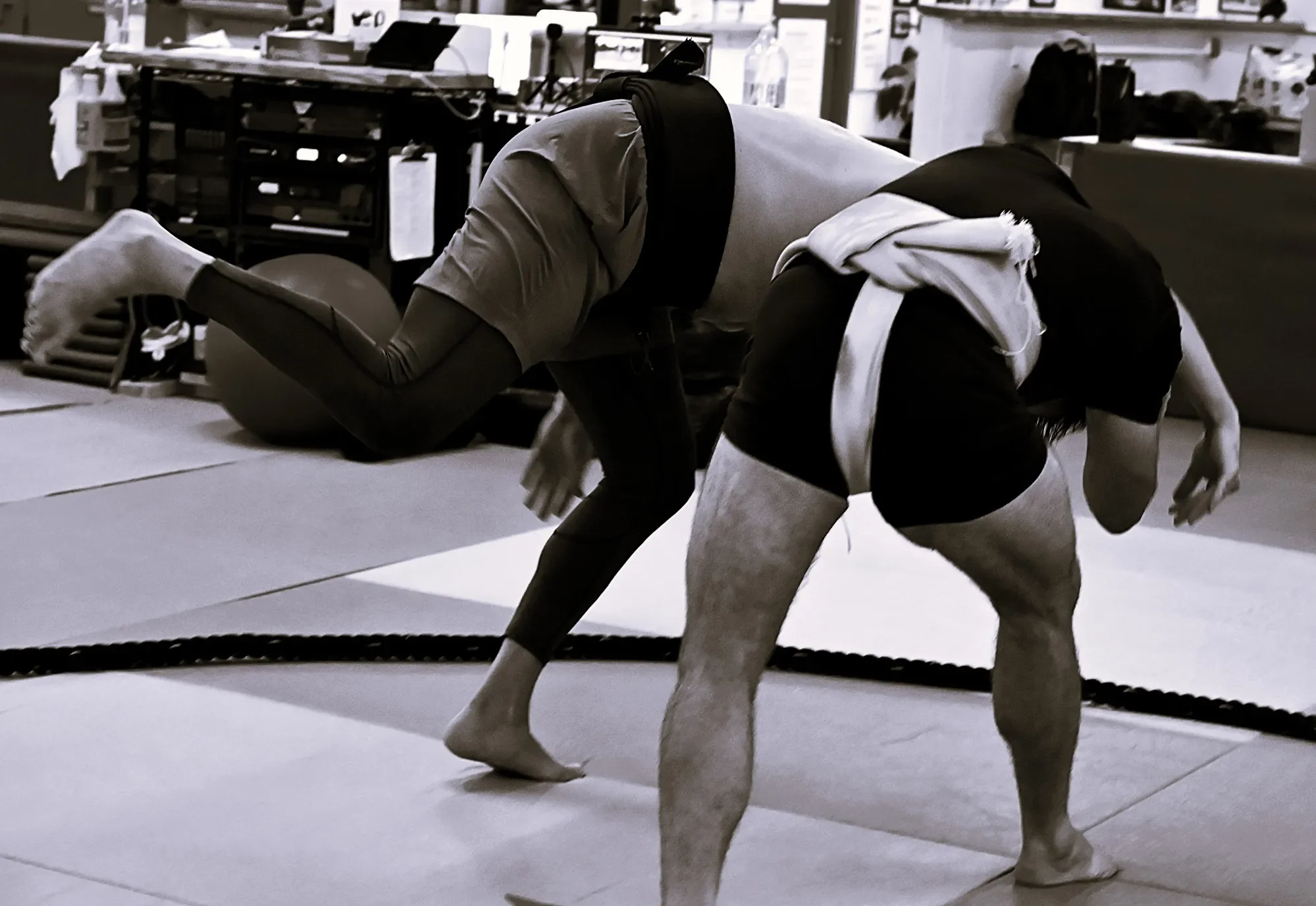 Two children practicing martial arts in a gym. One is wearing a black uniform and the other a white uniform. They are kneeling on mats, engaging in training.