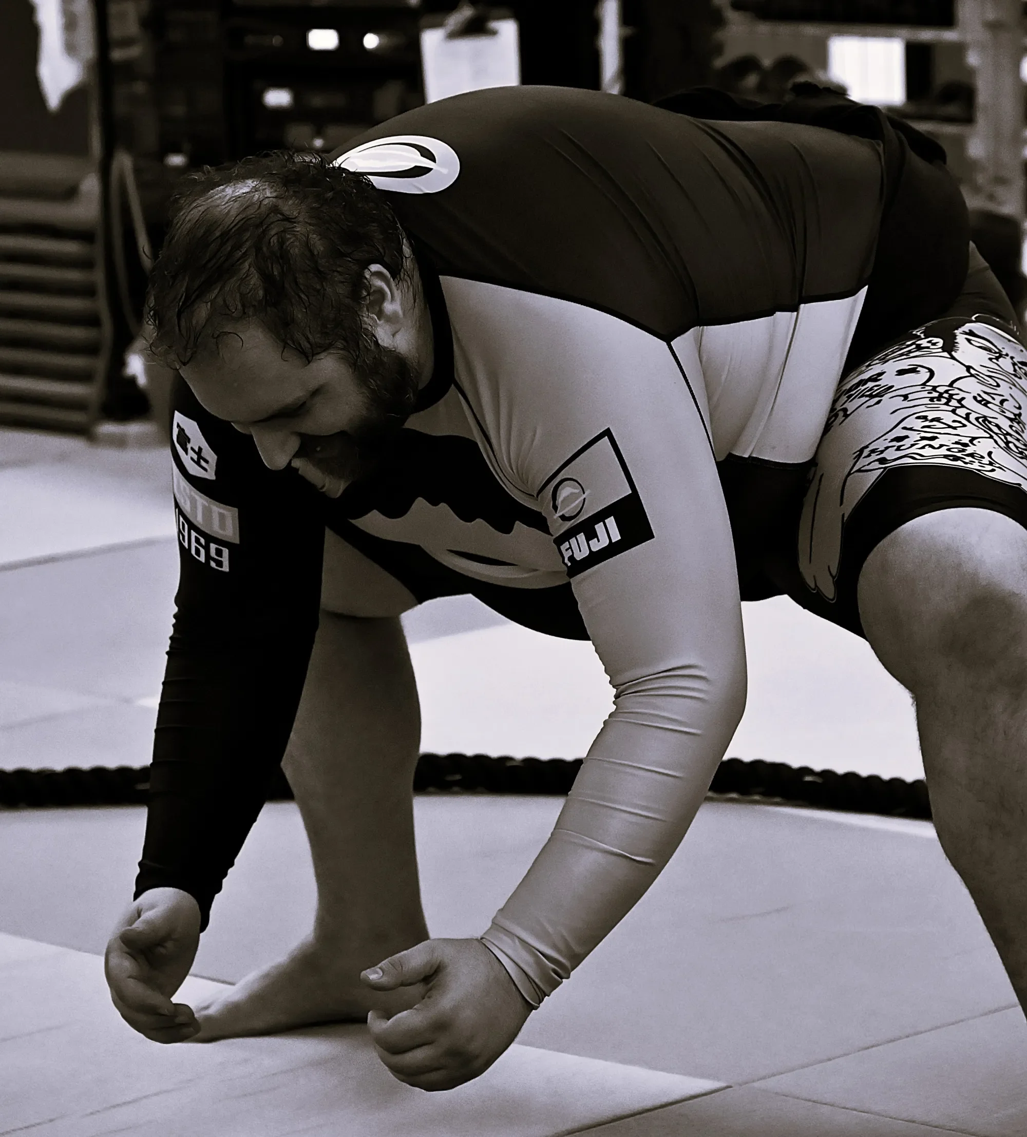 A fighter in a black and white sports uniform is on the ground, gripping the floor with one hand and bent over with their head down, during a wrestling match.