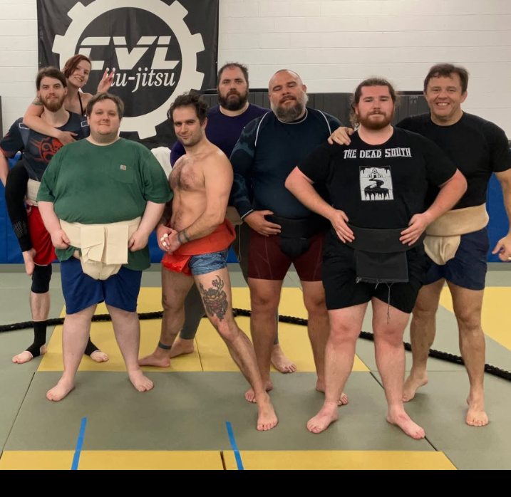 Group of eight people at a Brazilian Jiu-Jitsu gym, standing on mats in front of a black banner with the FVI FIGHT logo. They are dressed in martial arts attire, some wearing rash guards and shorts, others shirtless with gi pants or shorts, and a few