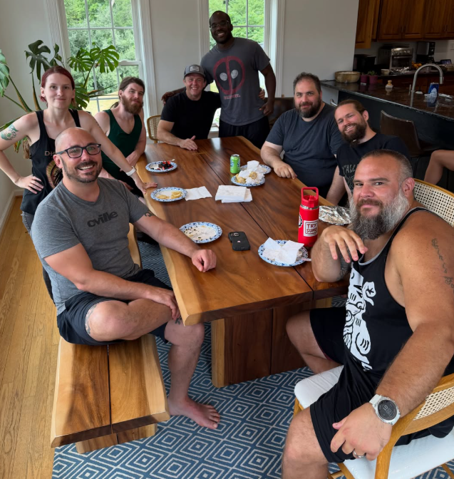 Group of eight people gathered around a wooden dining table in a bright kitchen, with some food and drinks, smiling and posing for the photo.
