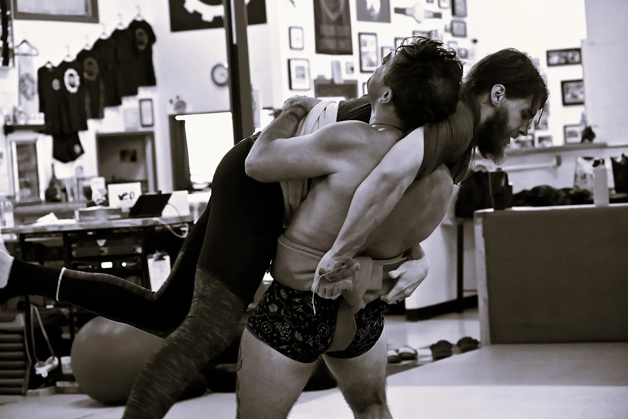 Two men wrestling on the floor in a room decorated with framed pictures and hanging shirts.