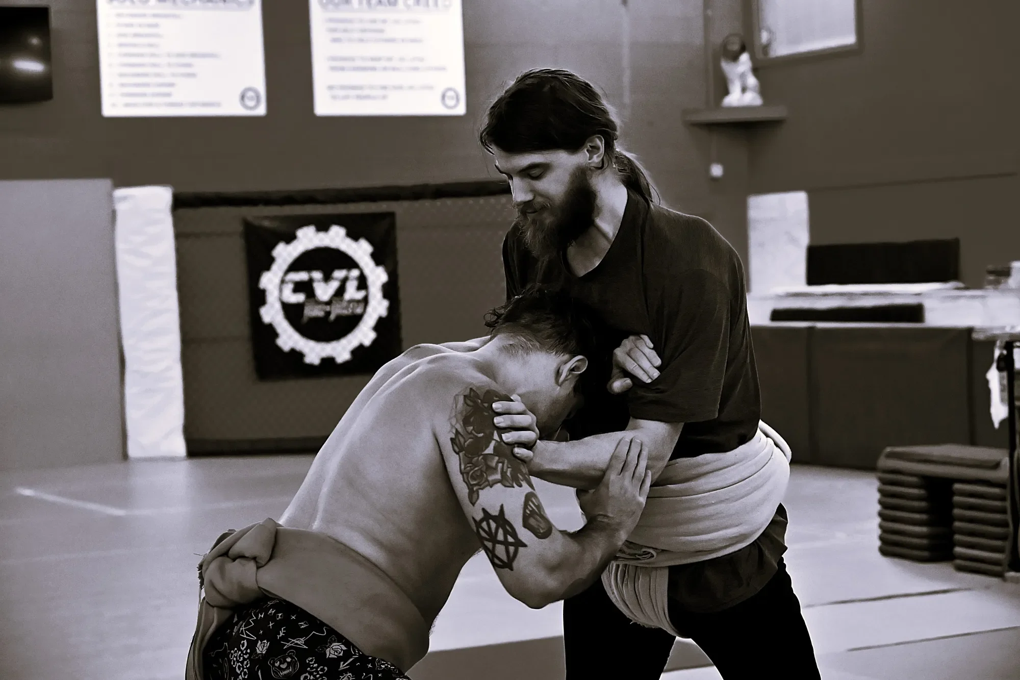 Two men practicing Brazilian Jiu-Jitsu in a gym, one man with tattoos grips the other man's arm while the other man presses him down on the mat, both are shirtless.