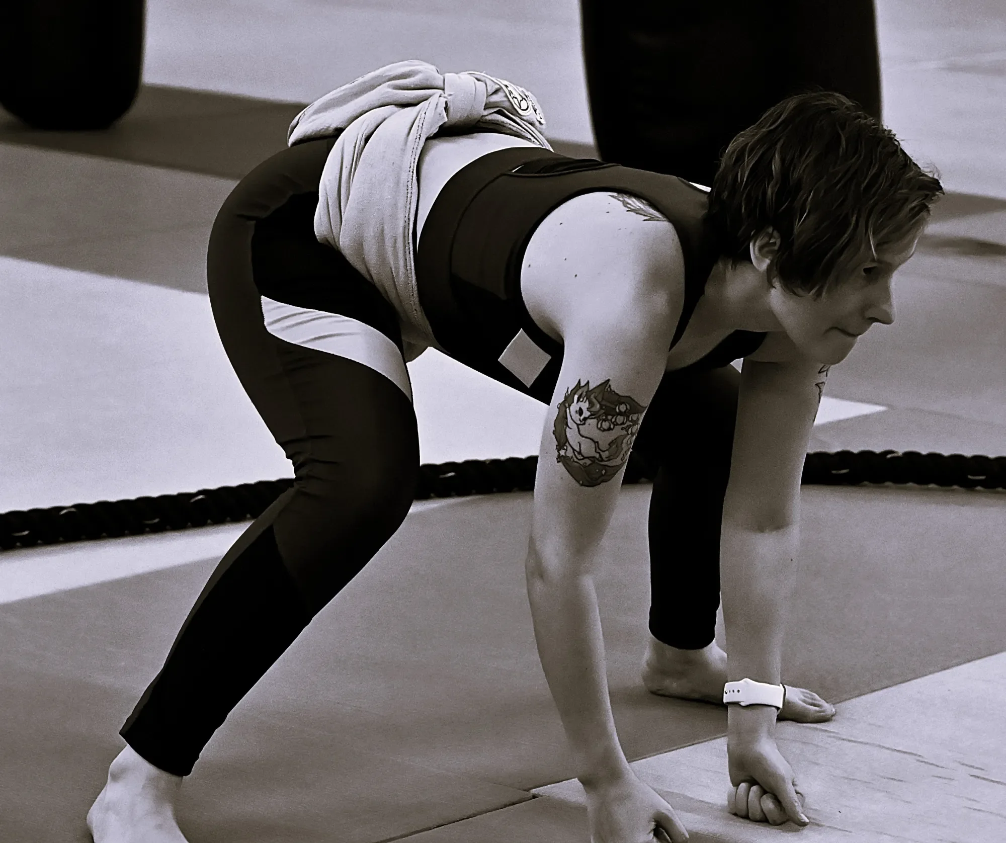 A woman is in a crouching position on a gray floor, wearing a sports outfit with a shirt and leggings, with one arm extended downward and her head lowered.