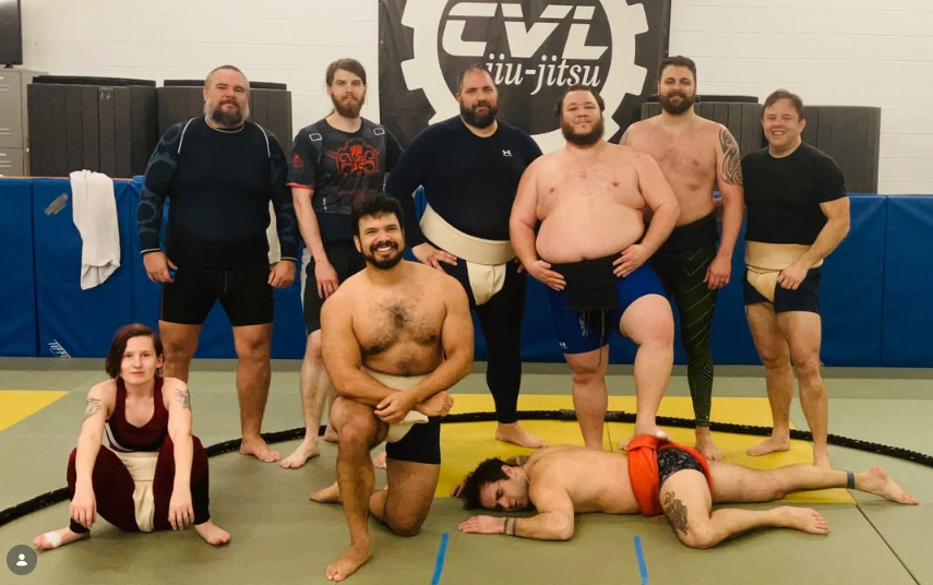 Group of seven men and one young girl standing or lying on a martial arts training mat in a gym, with a logo that says 'CVL jiu jitsu' on the wall behind them.