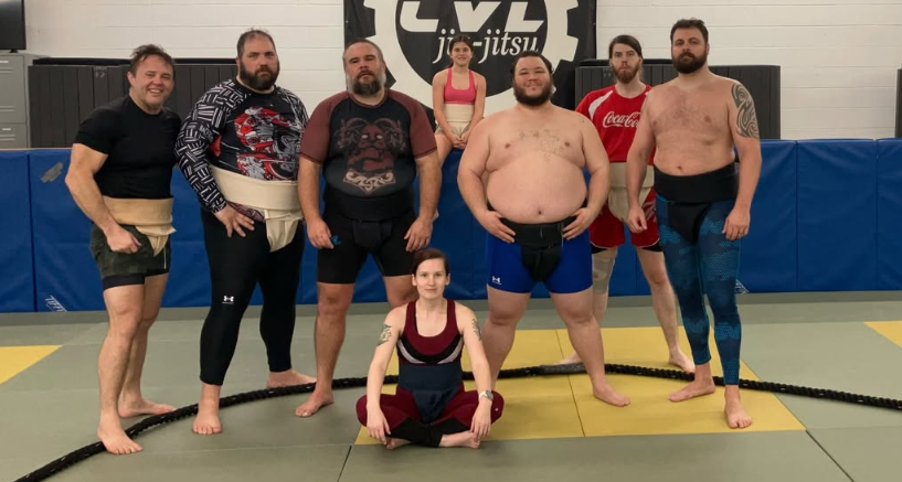 Group of people in martial arts uniforms and casual attire posing in a gym with a Jiu-Jitsu sign on the wall.