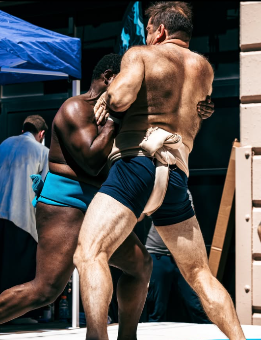 Two sumo wrestlers engaged in a match, wearing traditional mawashi loincloths, on a sumo ring. The match is intense, with one wrestler gripping the other's arm.
