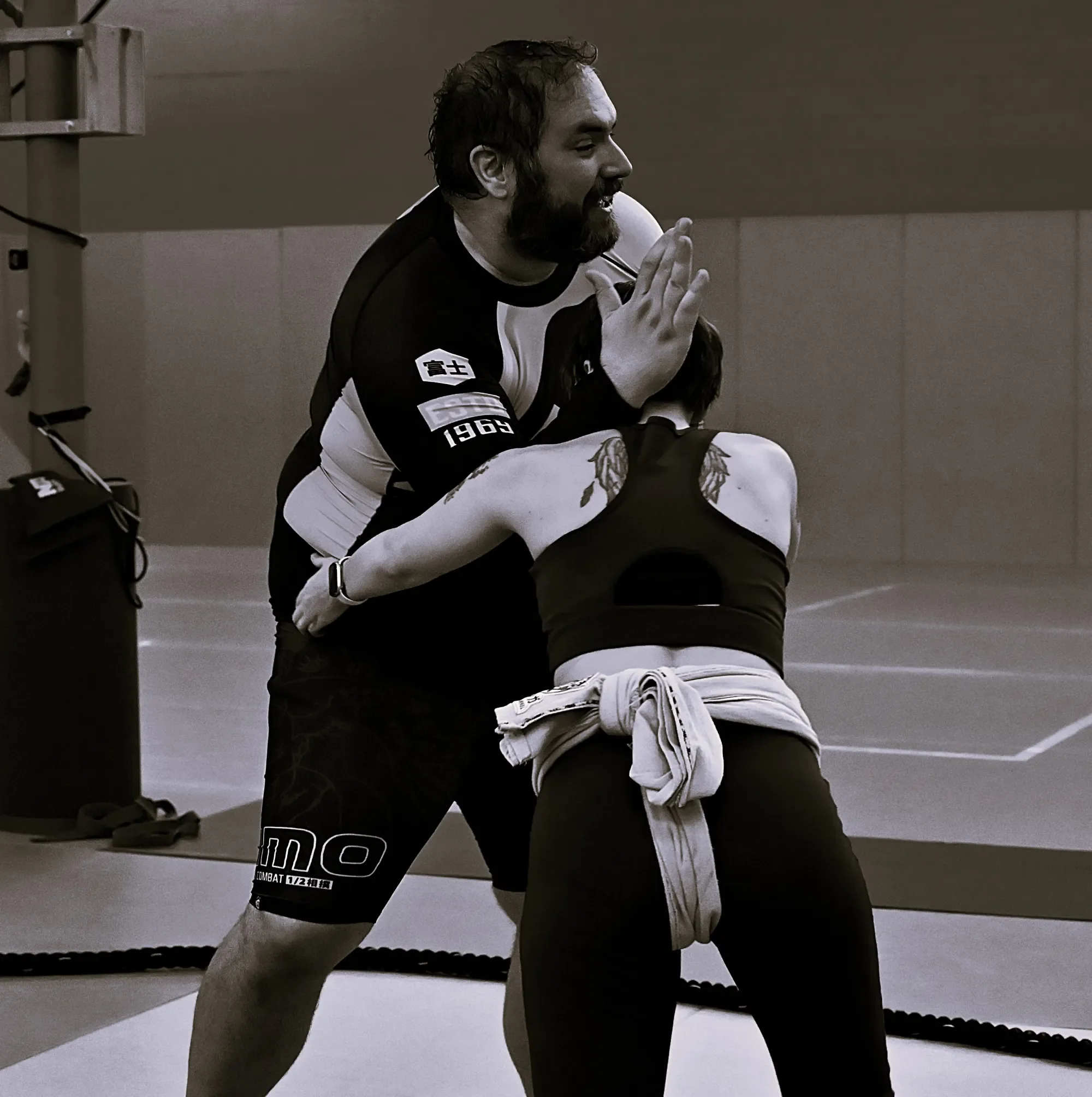 A man in a black and white sports uniform with the number 1963 on his arm, sitting on the floor and clasping his hands together in a prayer-like gesture, smiling with eyes closed in a room with a gray wall and gym equipment.