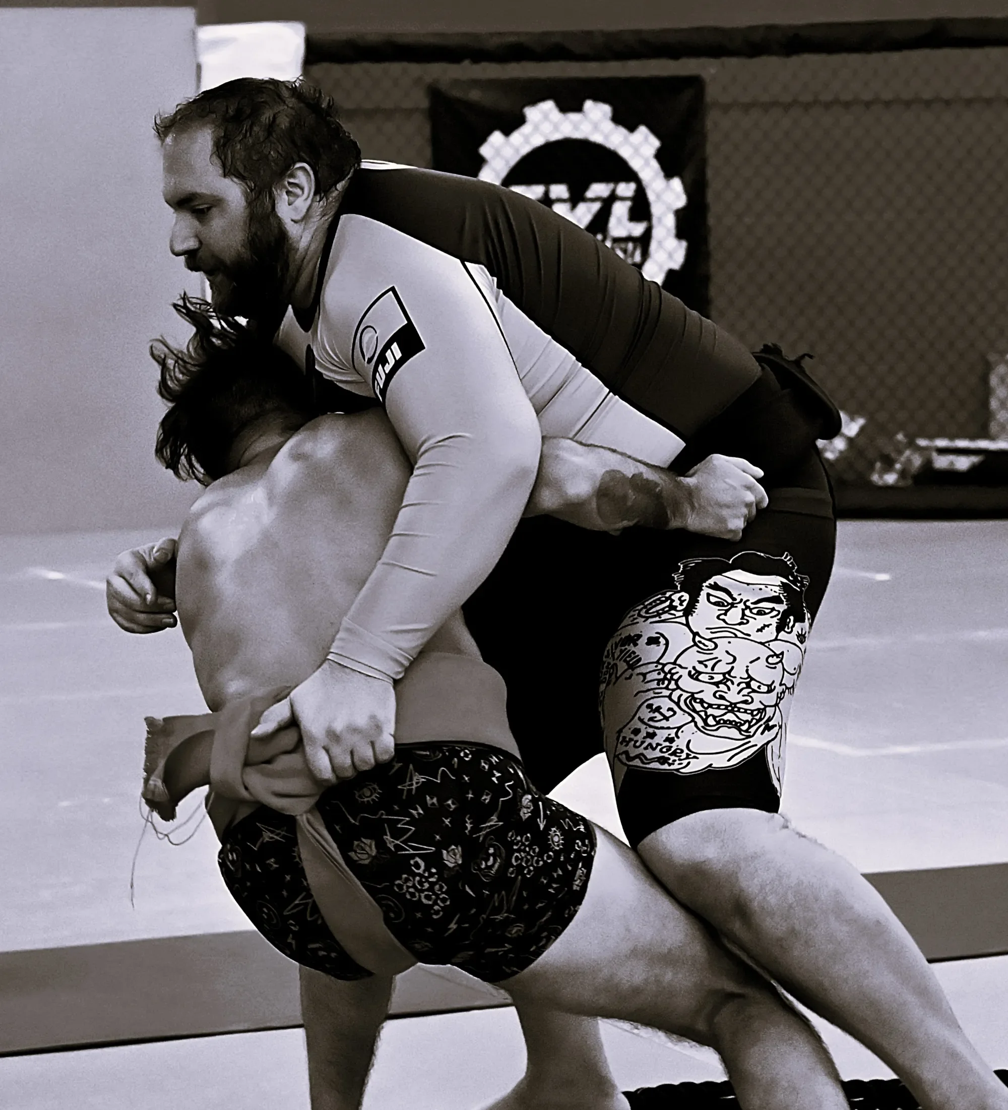 Two women practicing Brazilian Jiu-Jitsu on a mat, one in a white gi and the other in a black and colorful shorts, with a focus on their grappling hold and a cage wall in the background.