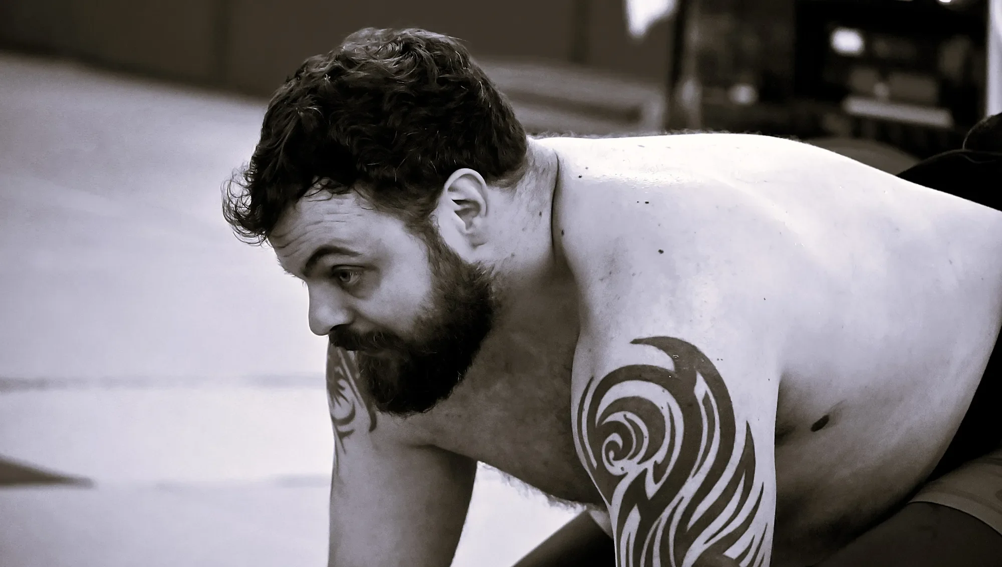 A shirtless young man with curly hair and a beard, lying face down with spiral tattoos on his shoulder and arm, in a gym or training environment.