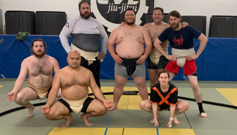 Six people, five men and one woman, posing in a martial arts gym with a jiu-jitsu sign behind them. Four men are standing, and two people are crouching in front. They are dressed in athletic gear, some with belts around their waists. The group appear