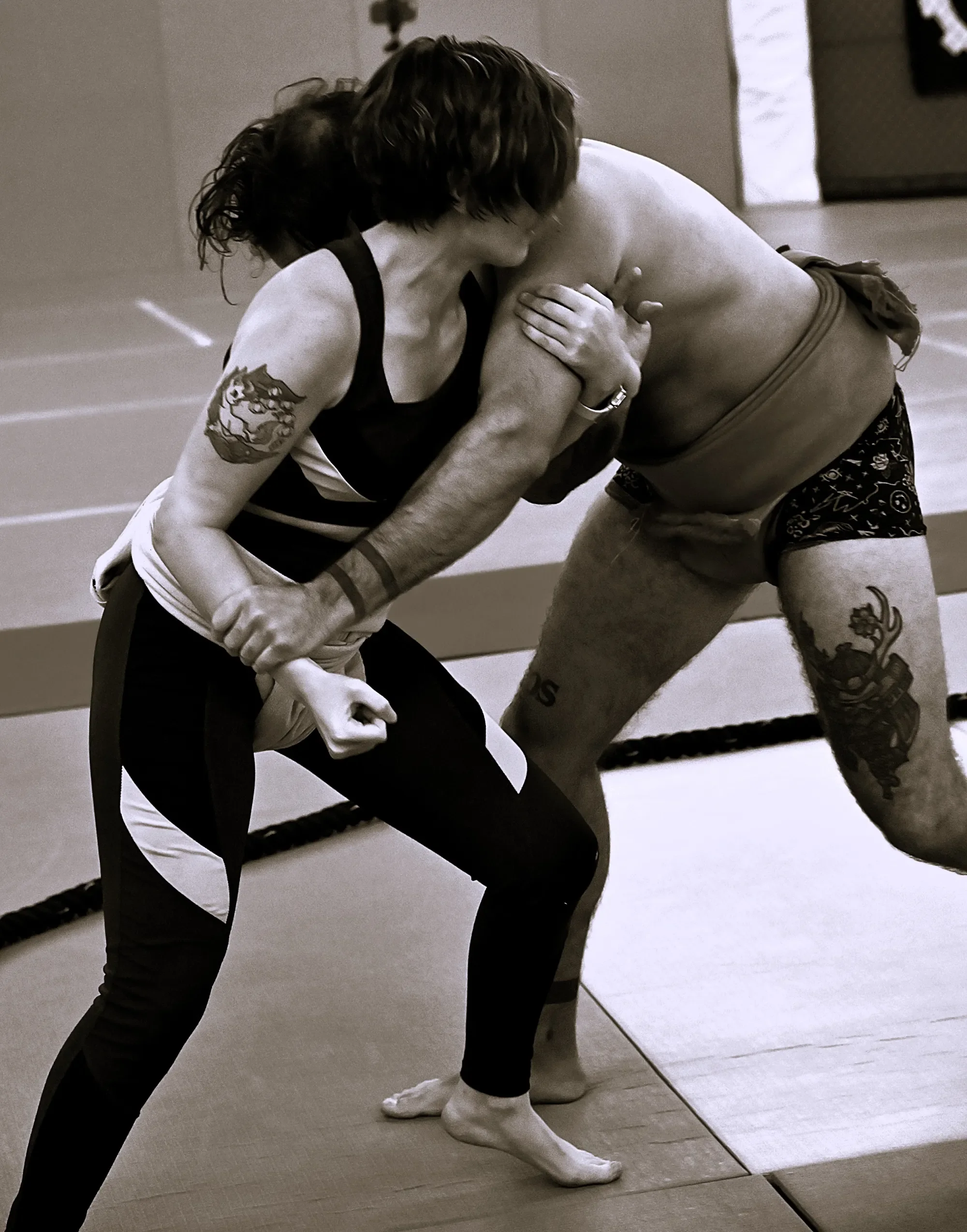 Two people practicing a grappling move in a martial arts dojo, one wearing a striped shirt with a tattoo on their arm and the other in shorts with a dragon tattoo, on a padded mat.
