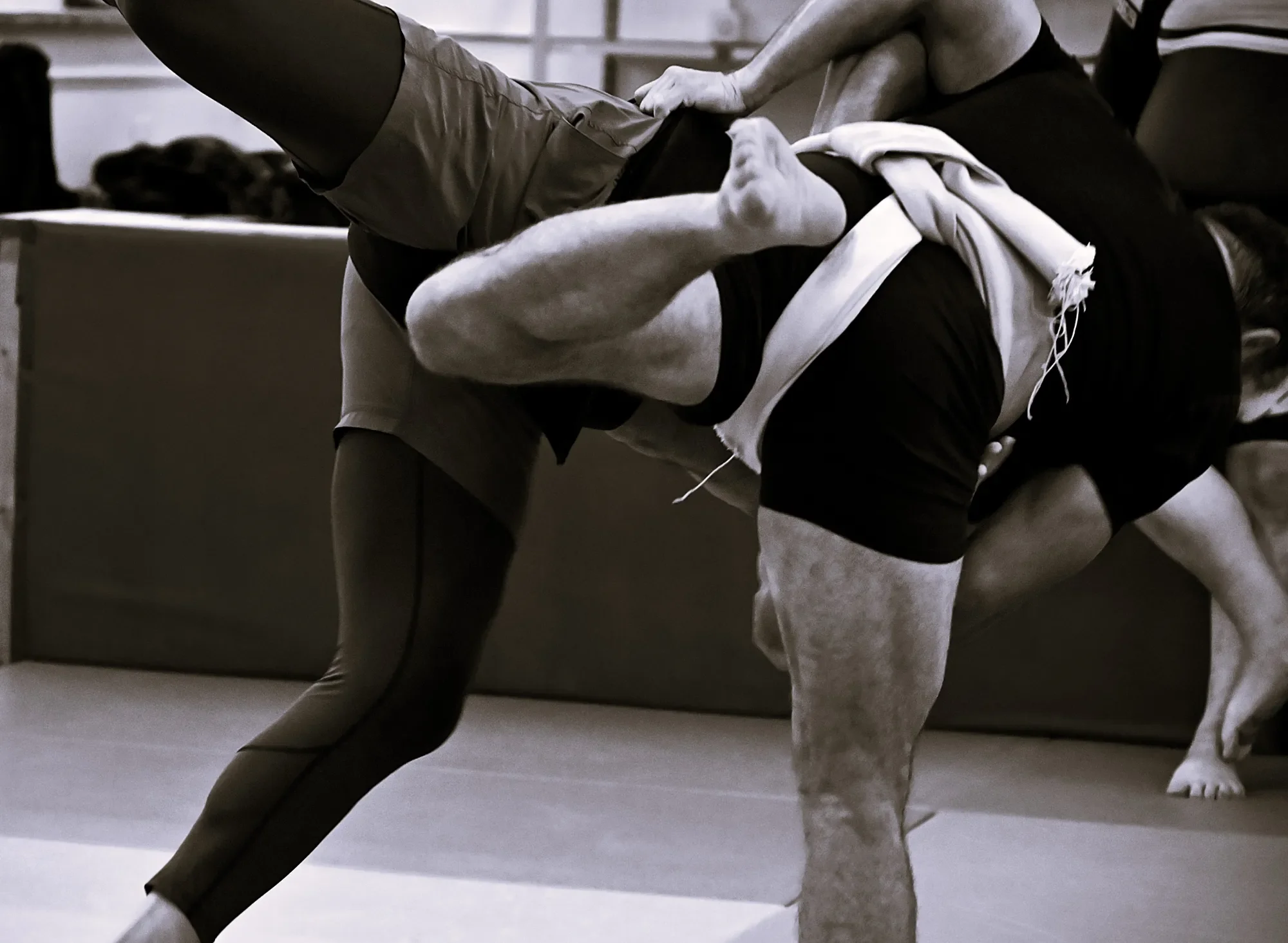 Two people practising martial arts, one a woman with a white gi, and the other a man with black attire, both engaged in a fight in a gym or dojo.
