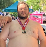 A man with a beard and short hair wearing a medal around his neck, shirtless, standing outdoors in front of colorful tents. It appears to be at an outdoor event or fair.