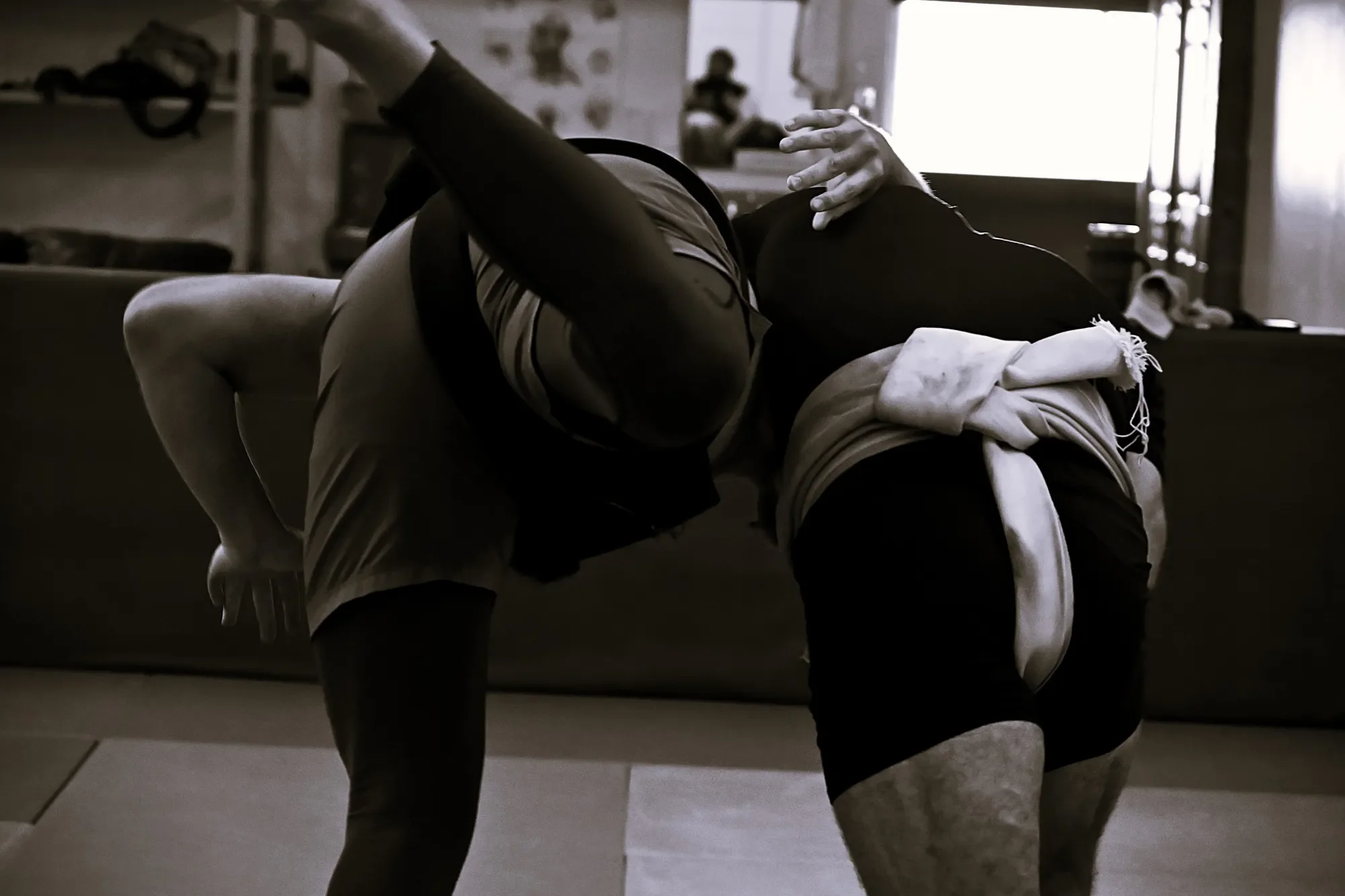 Two martial artists engage in a grappling match on a gym mat, with other practitioners and equipment visible in the background.