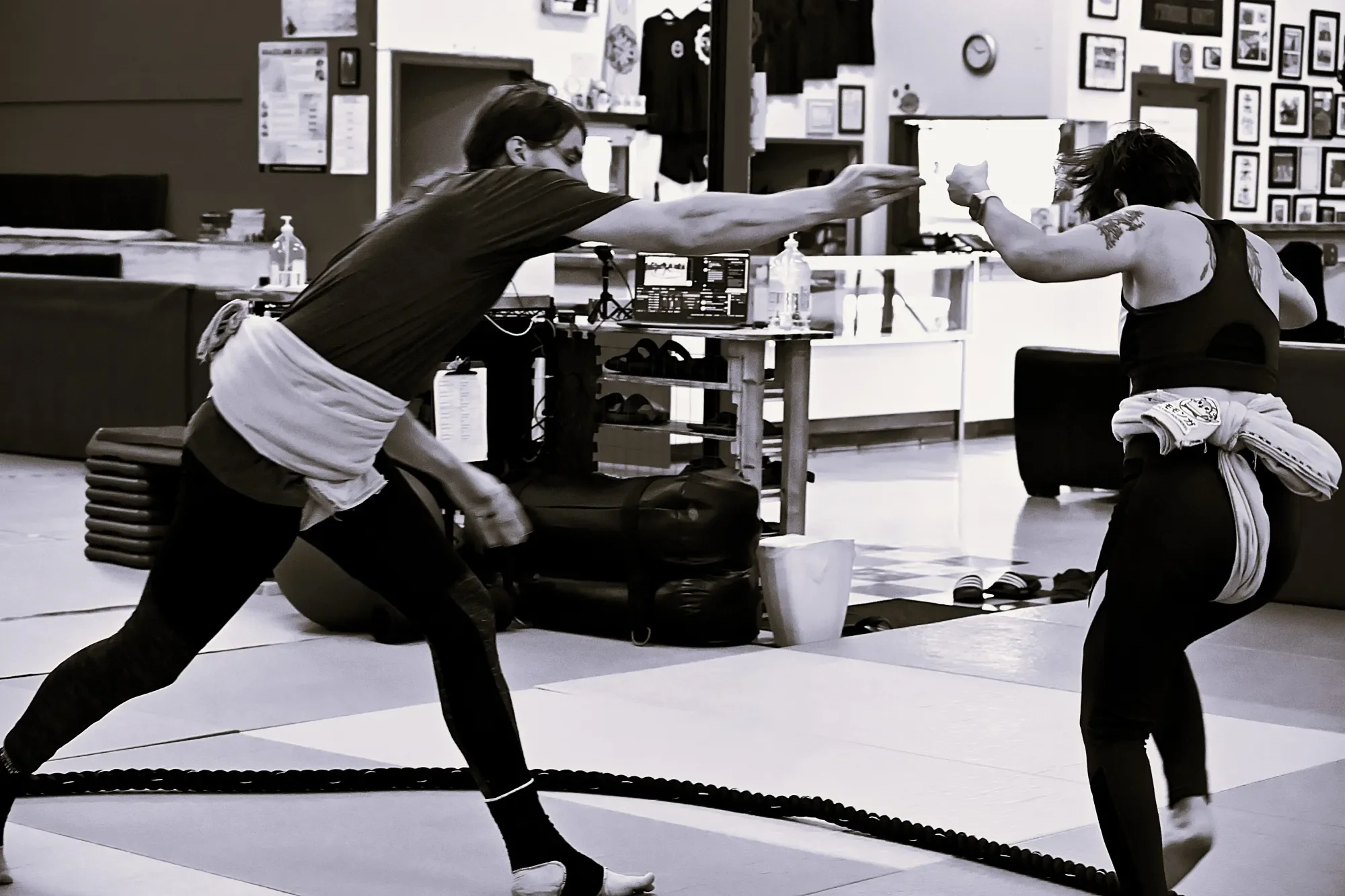Two women with tattoos practicing martial arts or self-defense in a martial arts studio with framed photos and artwork on the walls. One woman is reaching out her hand while the other is in a defensive stance, both wearing workout clothes.