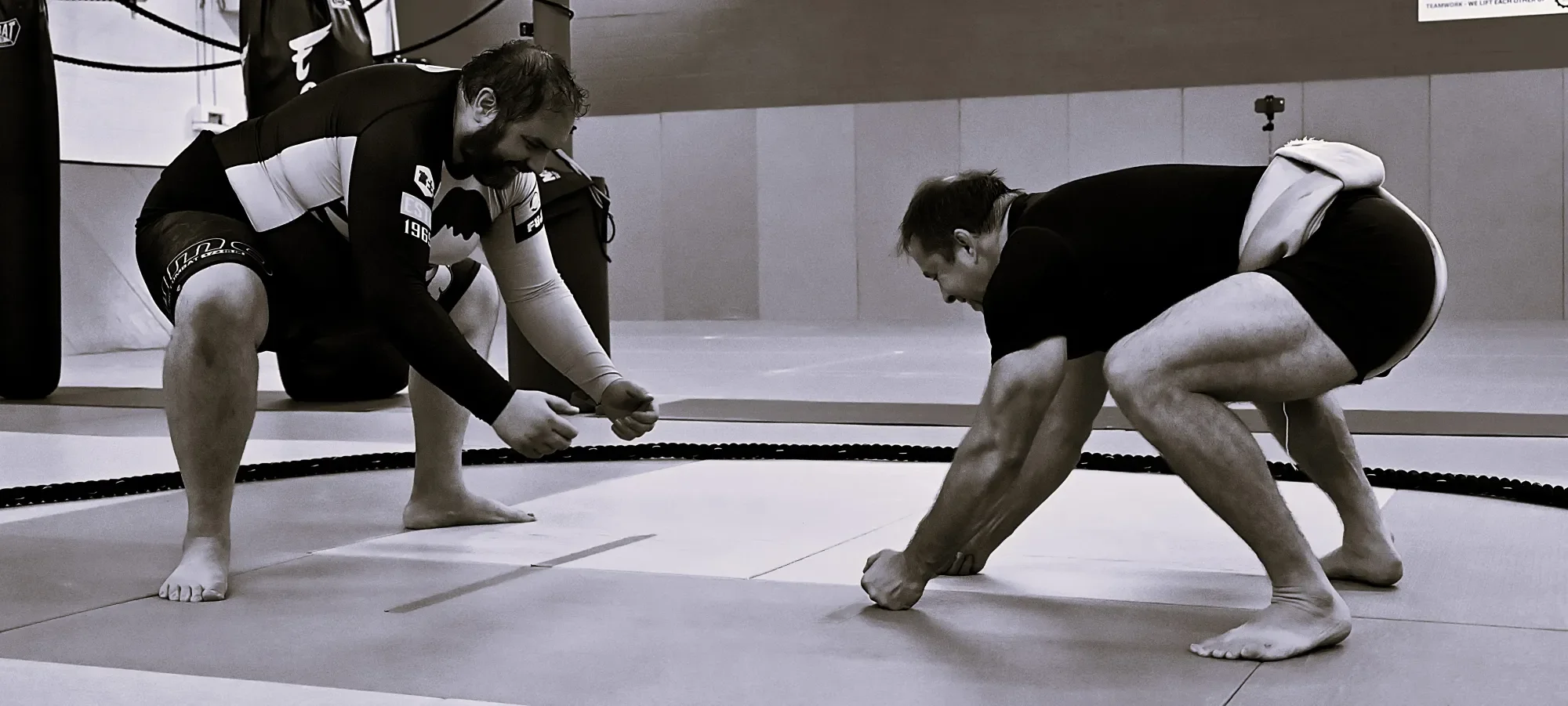 Two men in athletic clothing practicing Brazilian Jiu-Jitsu on grappling mats inside a gym, with one man performing a movement while the other evaluates.