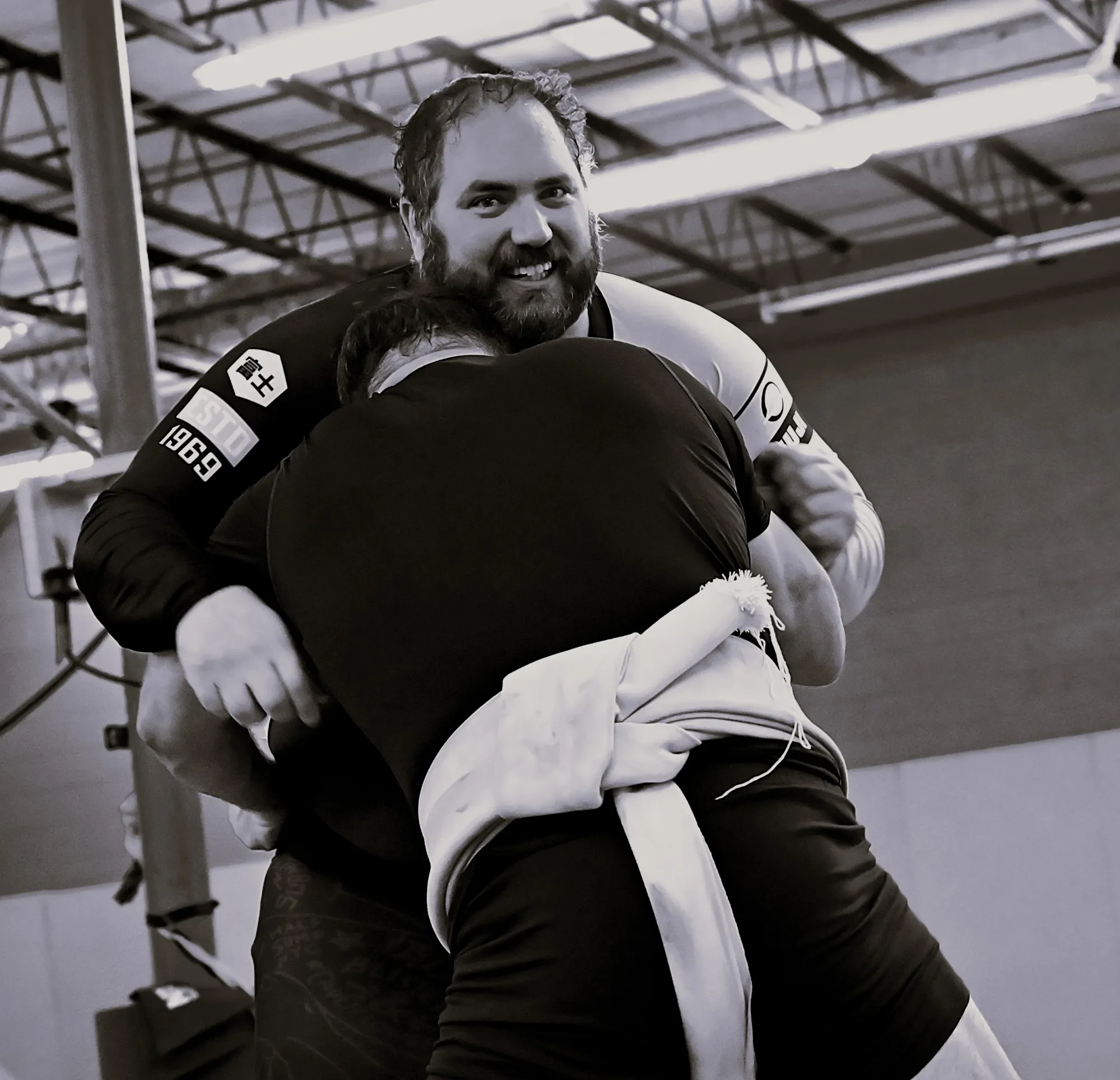 A man with a beard smiling, wearing a black and white sports uniform, hugging someone in a gym or sports facility.