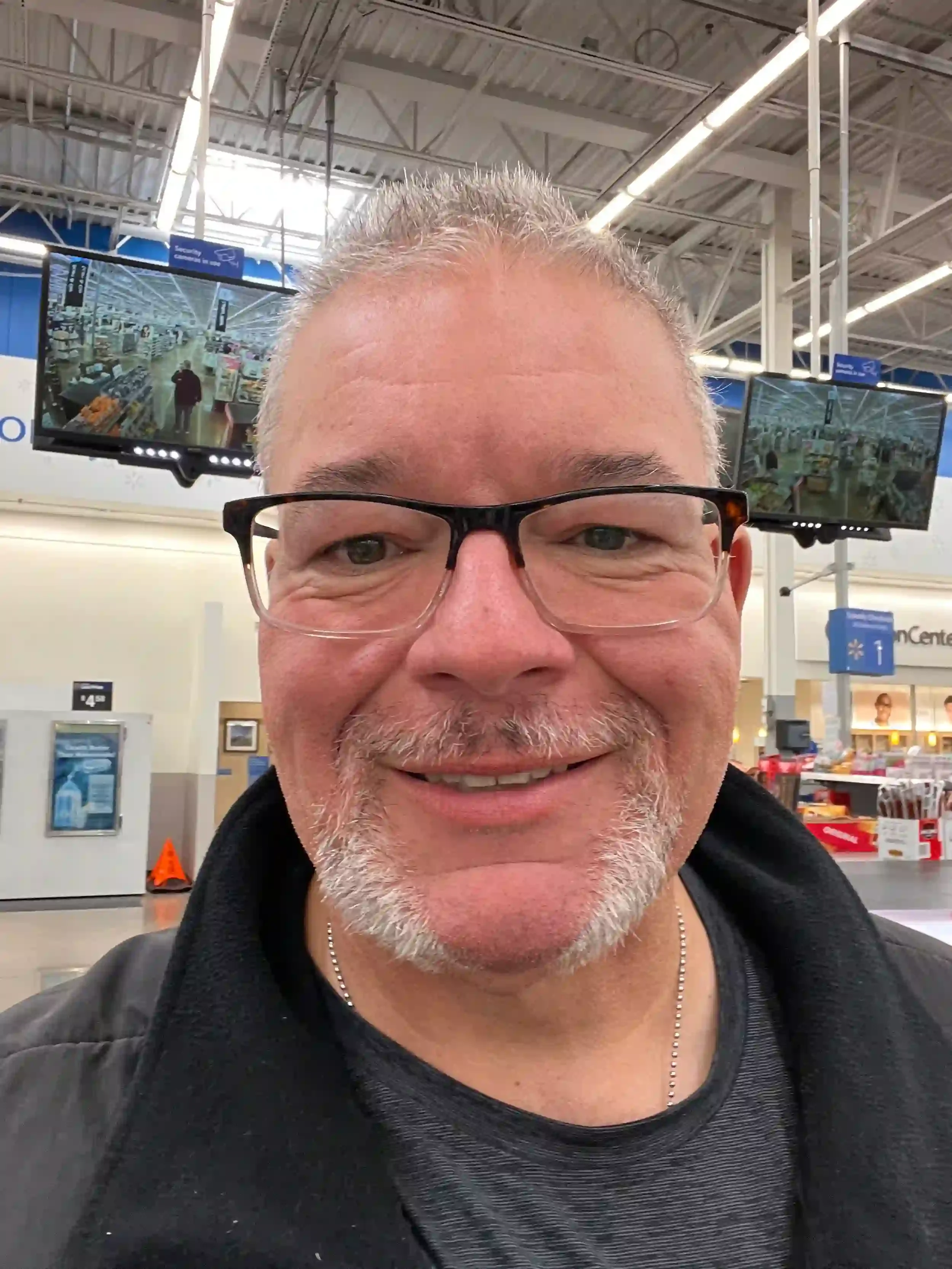 A man with glasses, a beard, and short gray hair taking a selfie in a store aisle, with multiple television screens behind him showing a store interior.