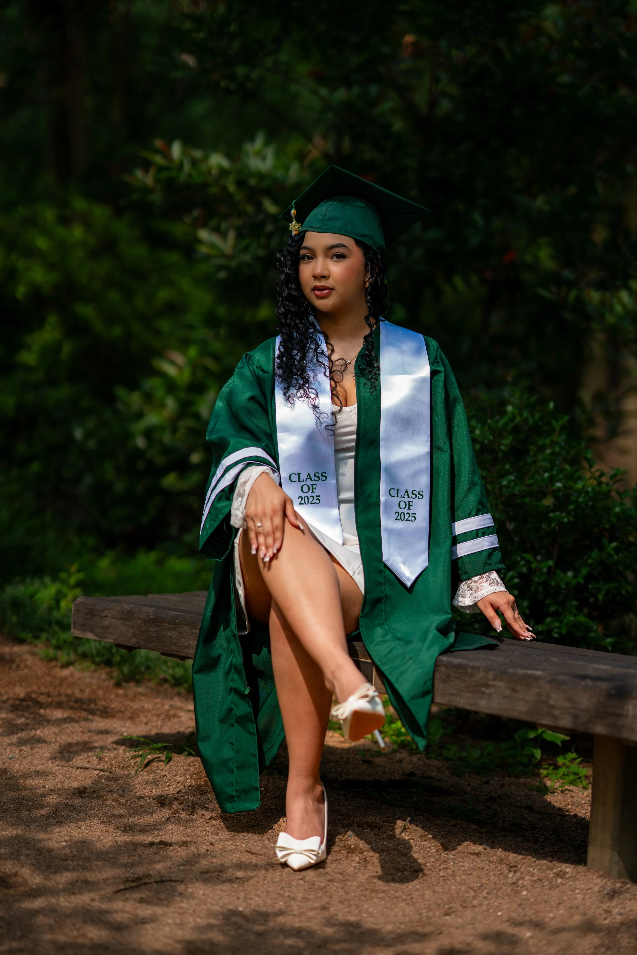 A young woman wearing a green graduation gown and cap, with a white stole that says "Class of 2025," sitting on a wooden bench outdoors surrounded by greenery. She has curly black hair, tan skin, and is wearing white high heels and lace sleeves.