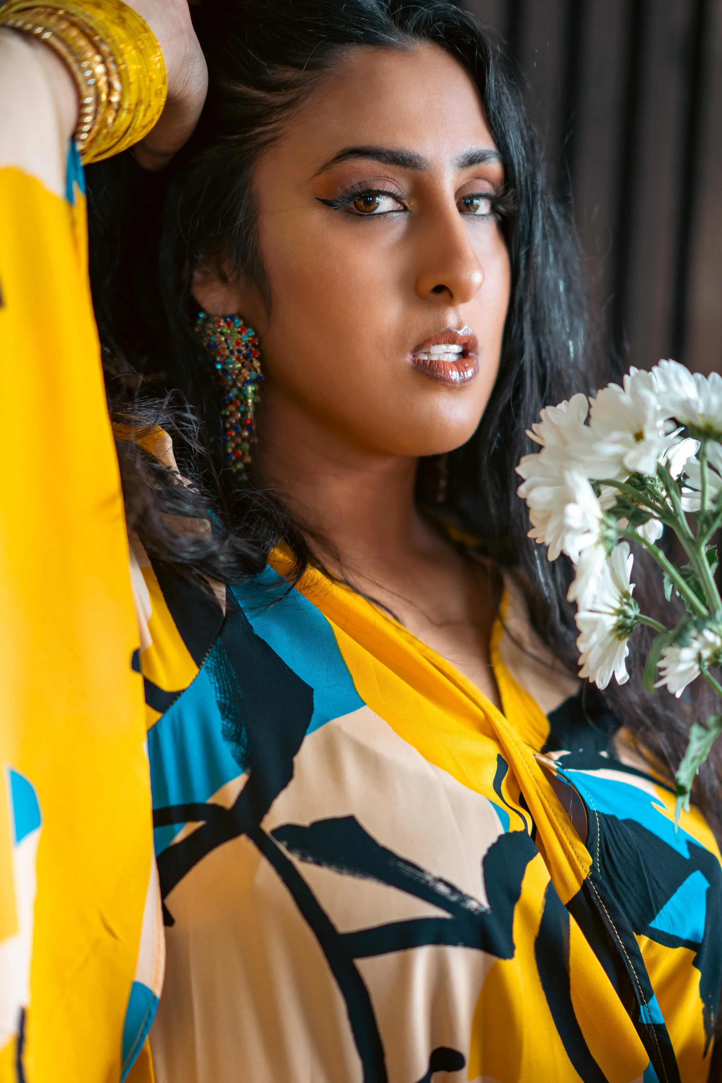 Close-up of a woman with dark hair, brown eyes, wearing colorful earrings, makeup, and a yellow, blue, black, and beige patterned jacket, holding white flowers near her face.
