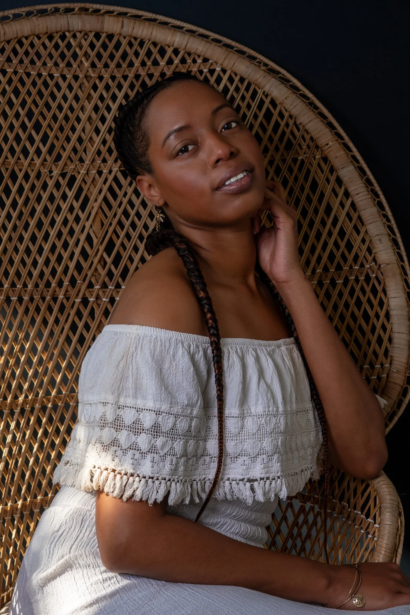 A woman sitting in a wicker chair, wearing a white off-the-shoulder top with lace details and a matching white skirt, against a dark background.