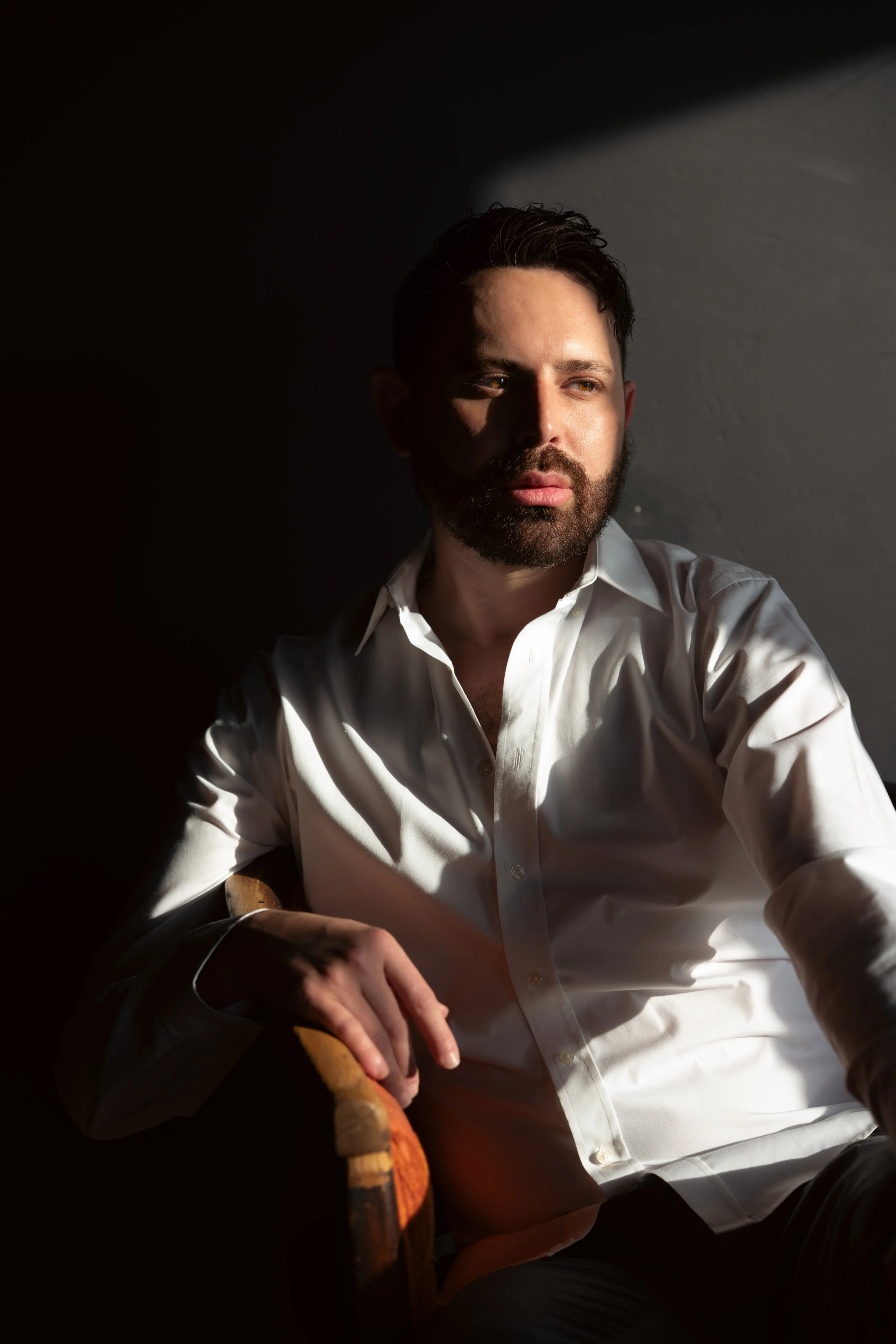A man with brown hair, a beard, and light skin, wearing a white button-up shirt, sitting on a chair with a wooden armrest, lit by sunlight coming from the right, with highlights on his face, dark background, and a neutral facial expression.