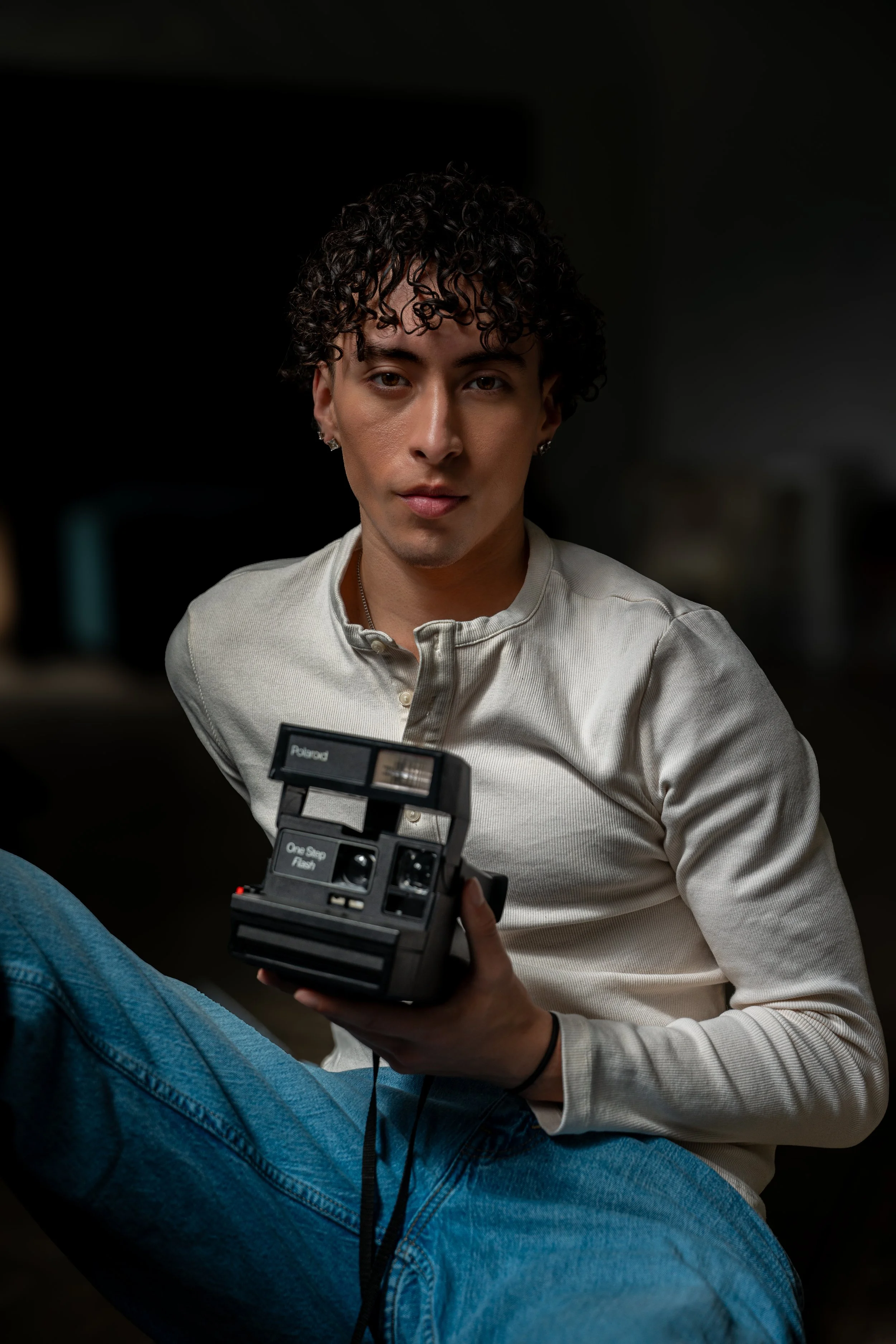 Portrait of a young man with curly hair wearing earrings and a cream-colored shirt, holding a Polaroid camera, sitting with crossed legs, and a person in blue jeans in the foreground.