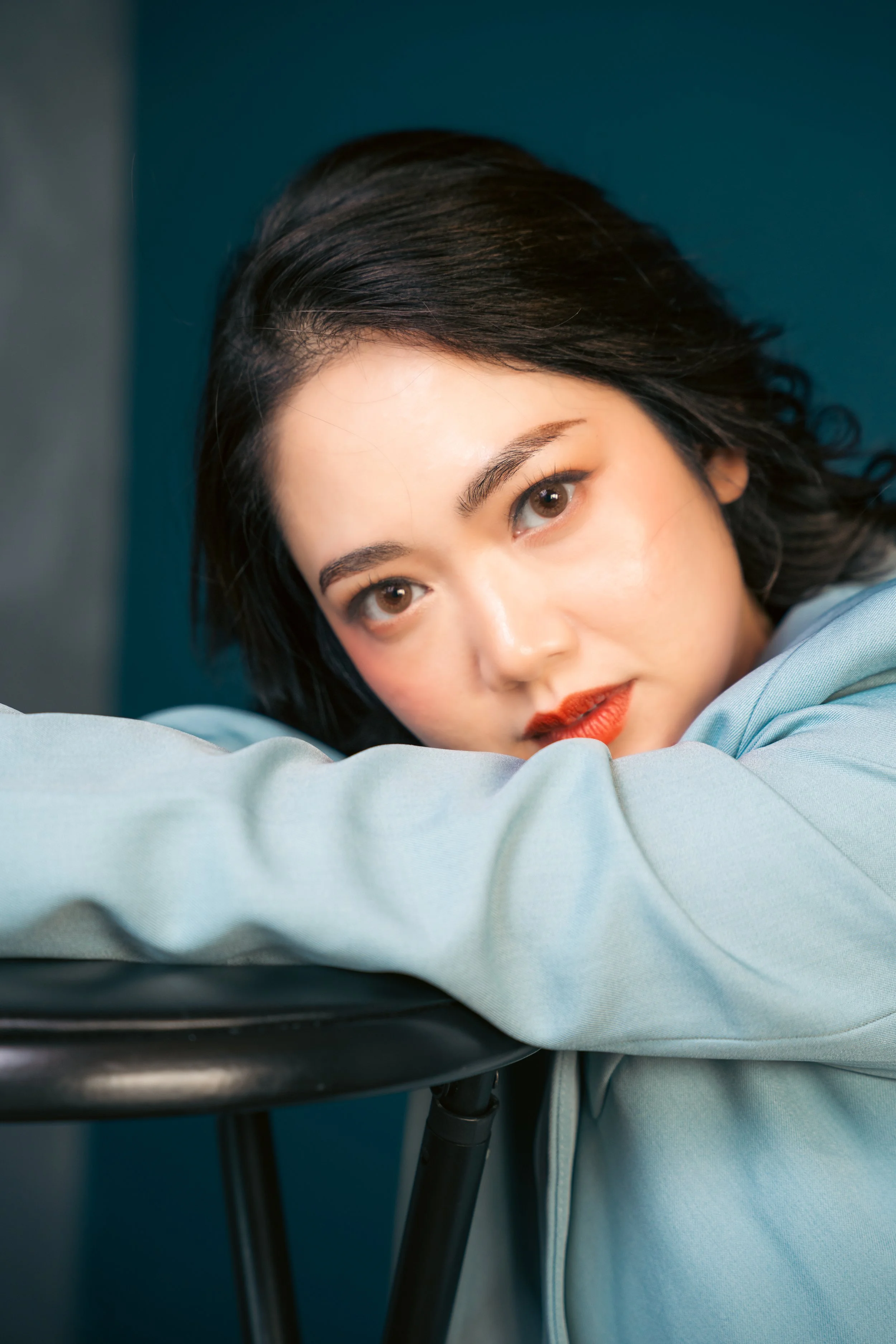 Close-up portrait of a young woman with dark hair lying on a light-colored surface, resting her head on her arm, with a blue background.