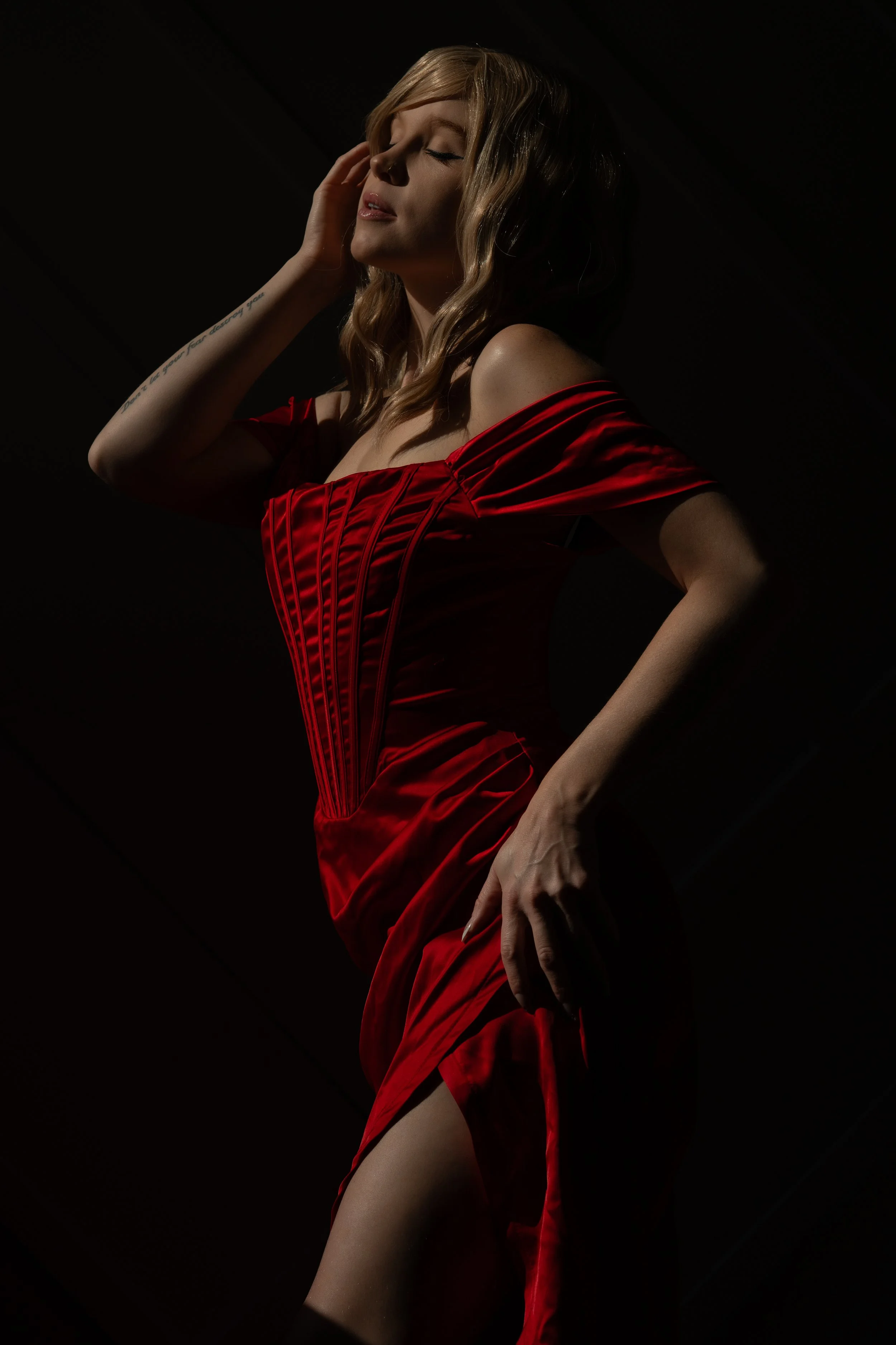 A woman in a red dress with corset-style details poses with her eyes closed and her left hand touching her face, against a dark background.