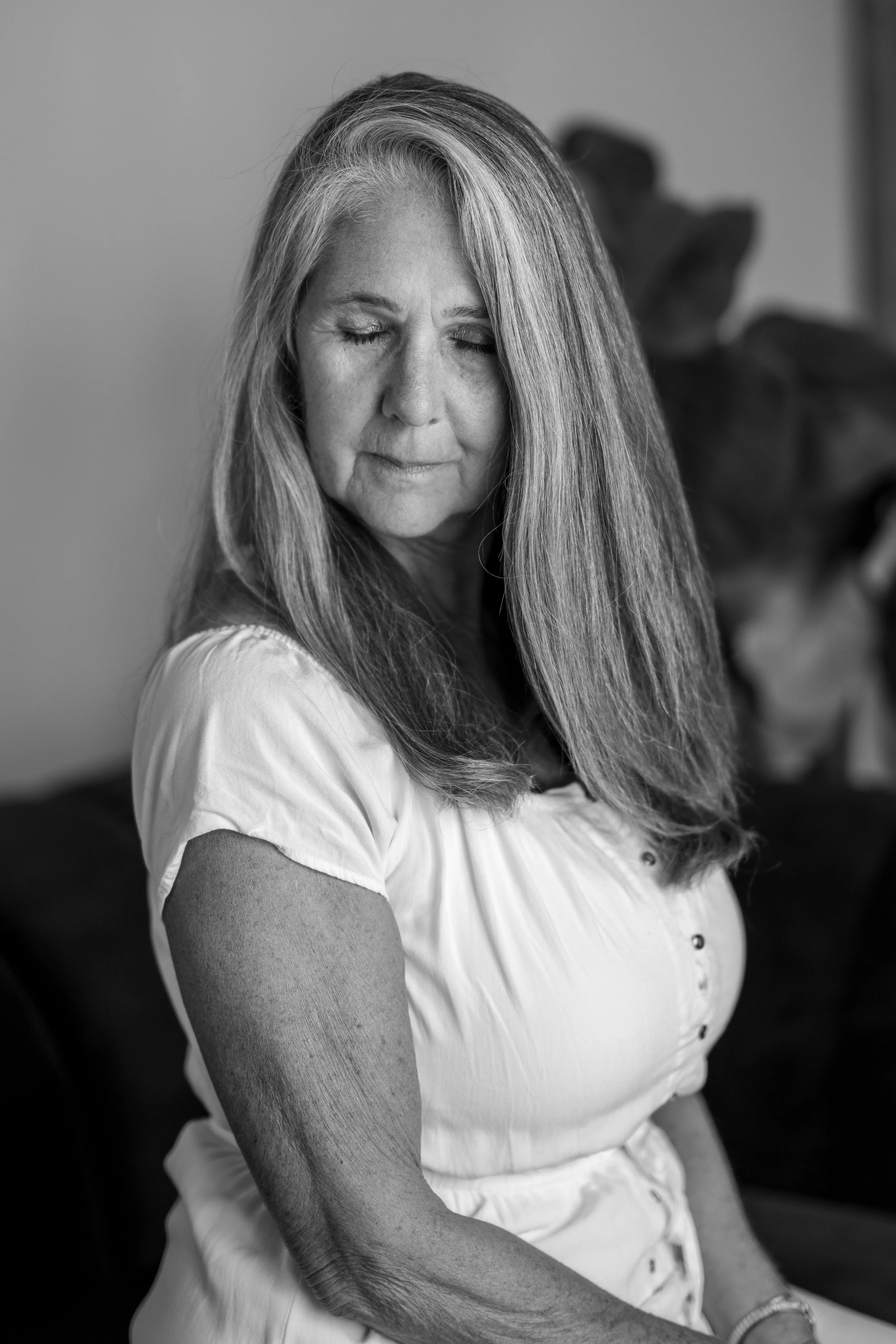 A black and white photo of an older woman with long hair, eyes closed, wearing a short-sleeved button-up shirt, and sitting indoors with a blurred plant in the background.