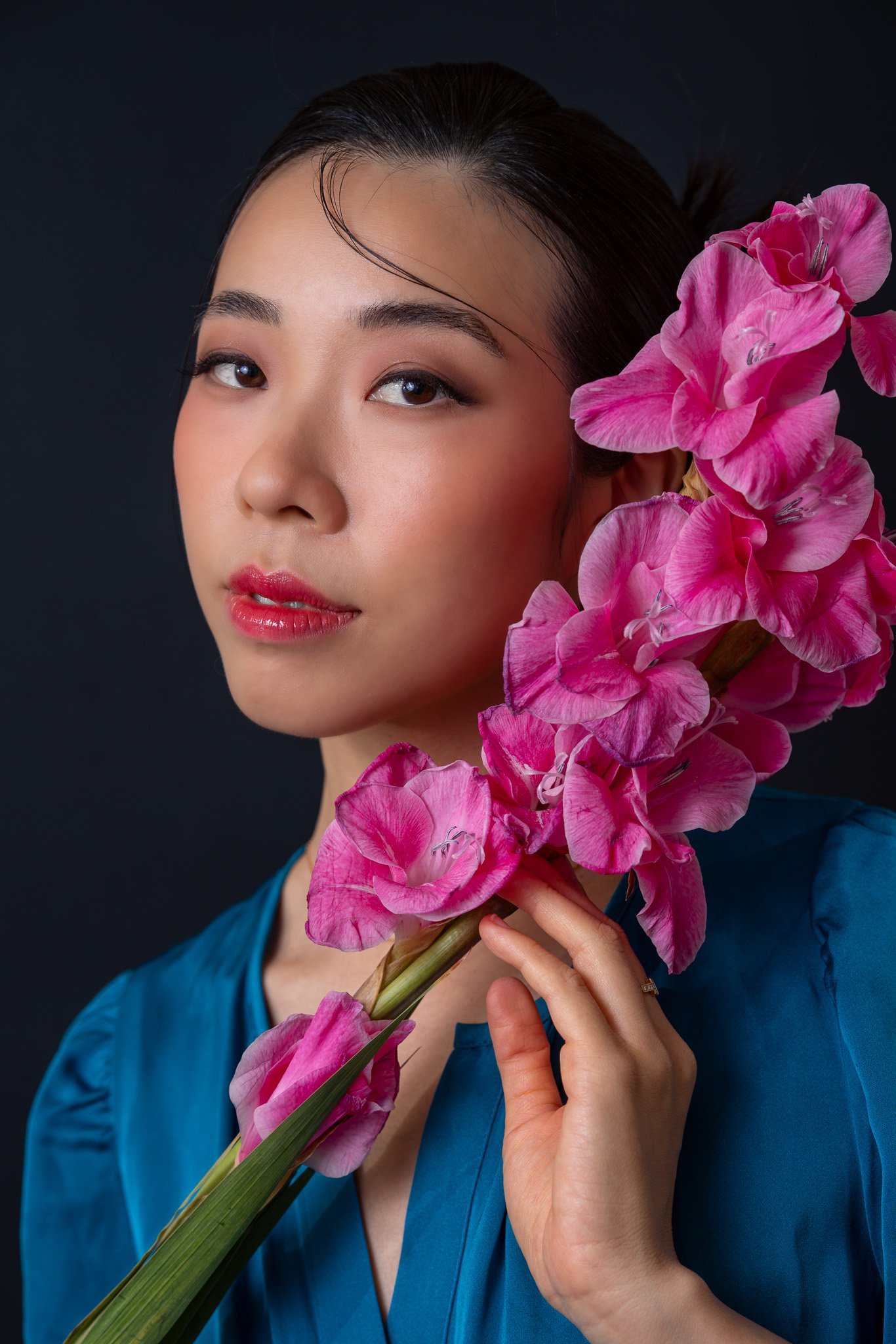 A woman with dark hair and makeup holding a bouquet of pink flowers against a black background.