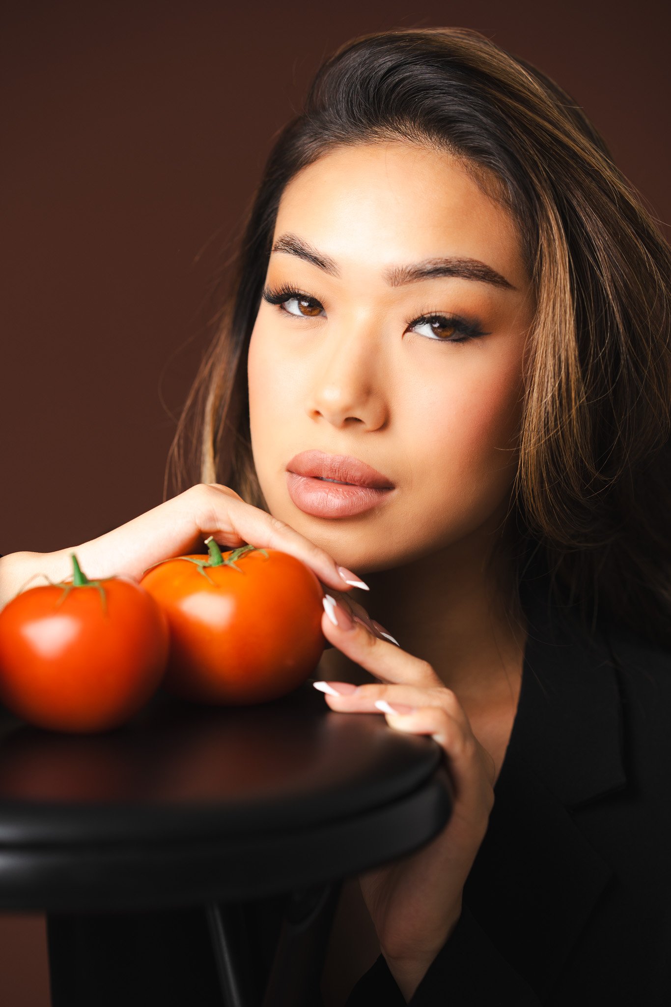 A woman with long, dark hair and makeup, looking at the camera while holding four ripe tomatoes in her hand with a dark background.