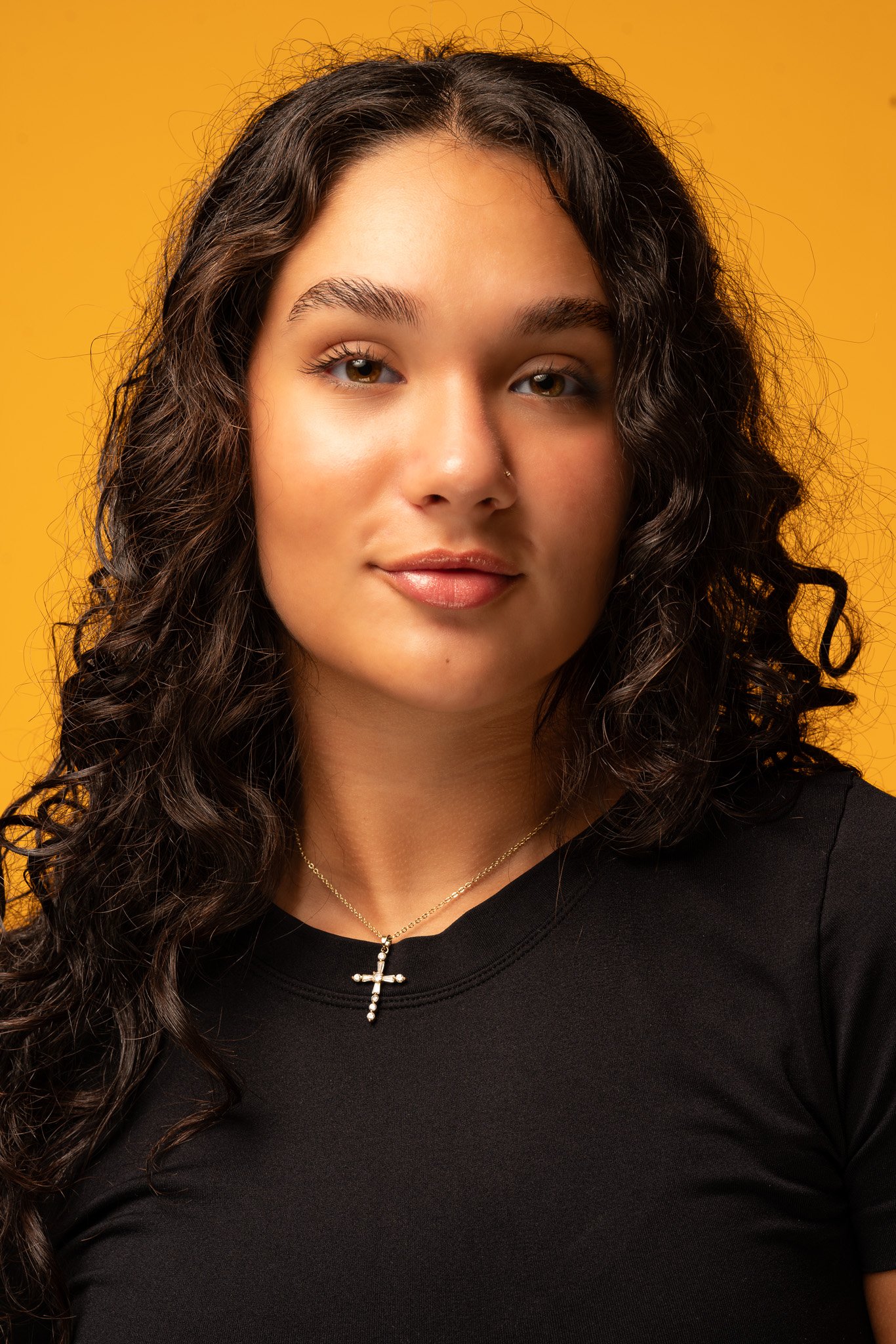 A woman with long, curly dark hair wearing a black top and a gold necklace with a cross pendant, standing against an orange background.