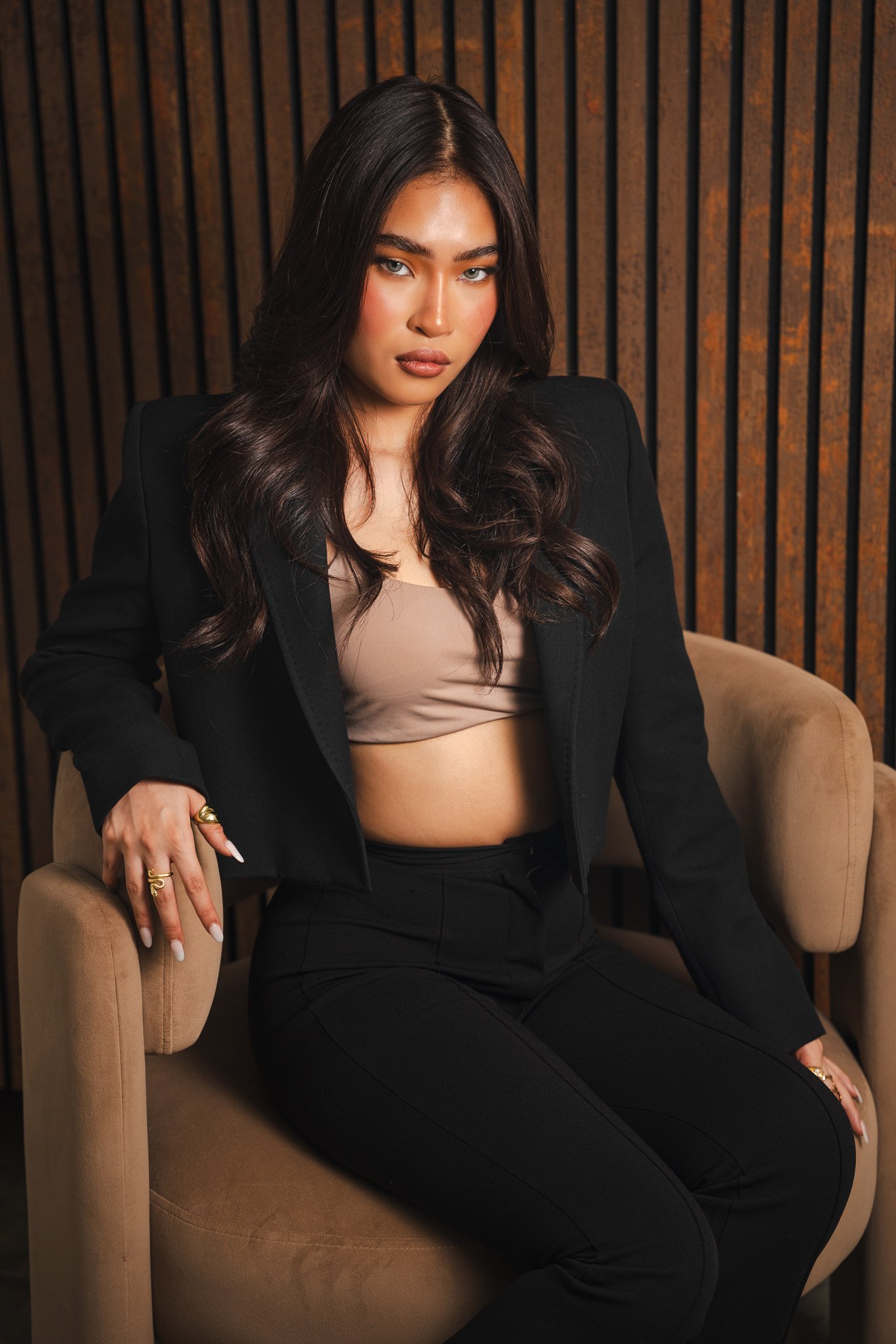 A woman with long dark hair and light skin, dressed in a beige crop top and a black blazer, is sitting on a beige chair against a wooden slatted background. She has a confident expression and positioned her left hand on the chair's armrest.