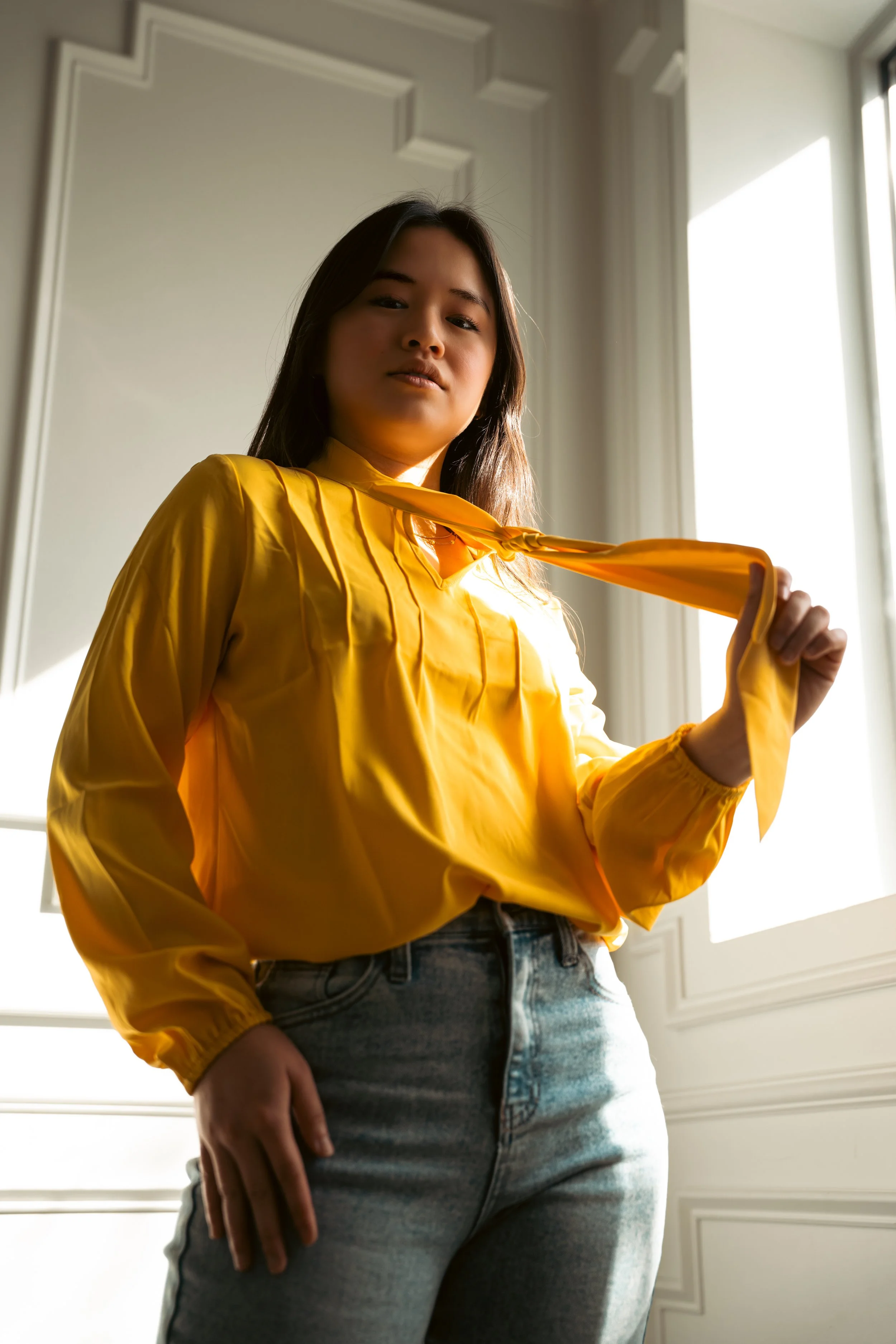 A woman wearing a yellow satin shirt and jeans standing inside near a window with white decorative wall panels, holding the collar of her shirt with her right hand, with natural light illuminating the scene.