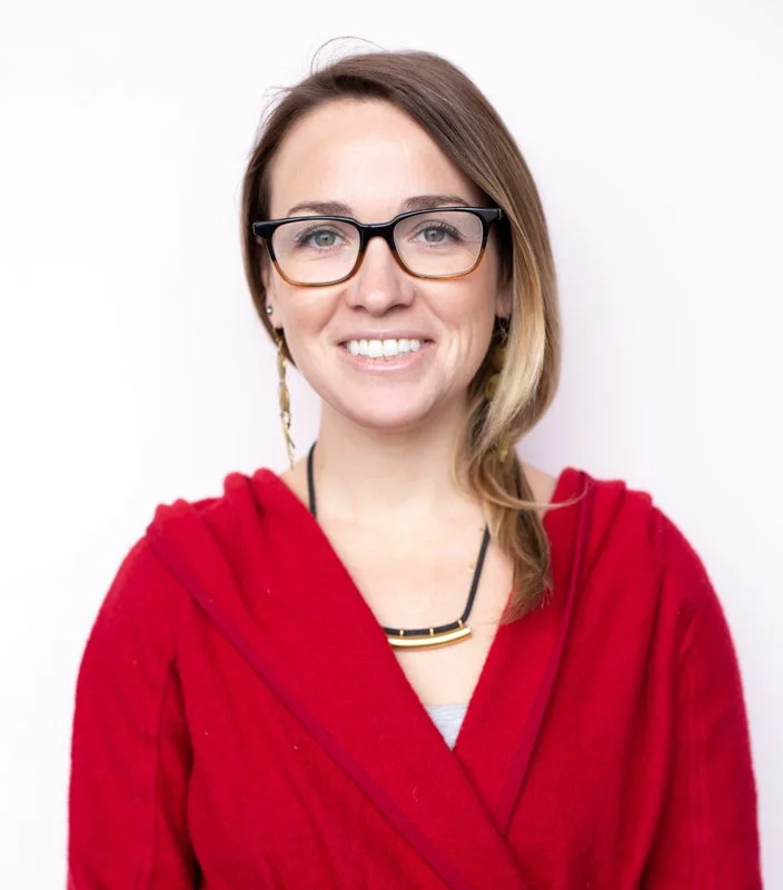 A smiling woman with shoulder-length light brown hair, wearing black-rimmed glasses, a red hoodie, a necklace, and earrings, against a plain white background.
