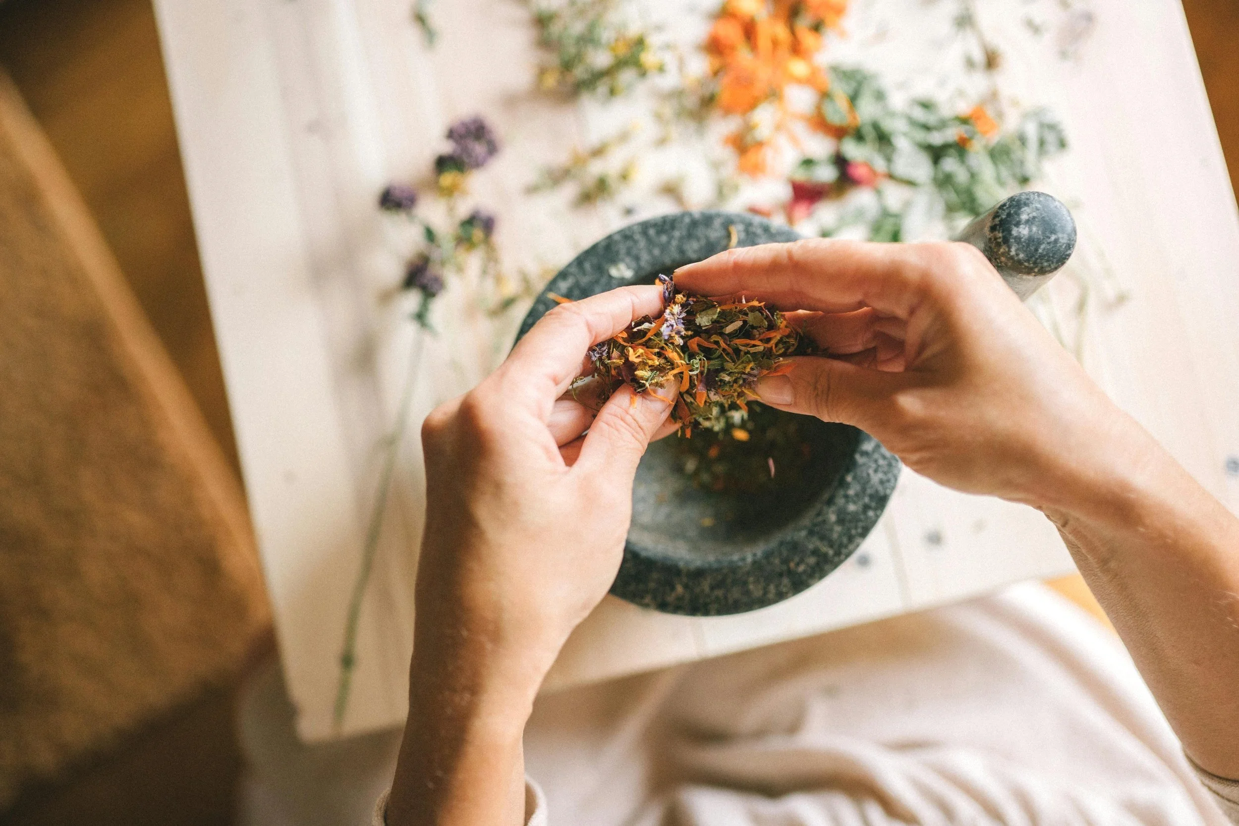 Hands grinding dried herbs using a mortar and pestle, with herbal remnants and flowers scattered on a white surface.