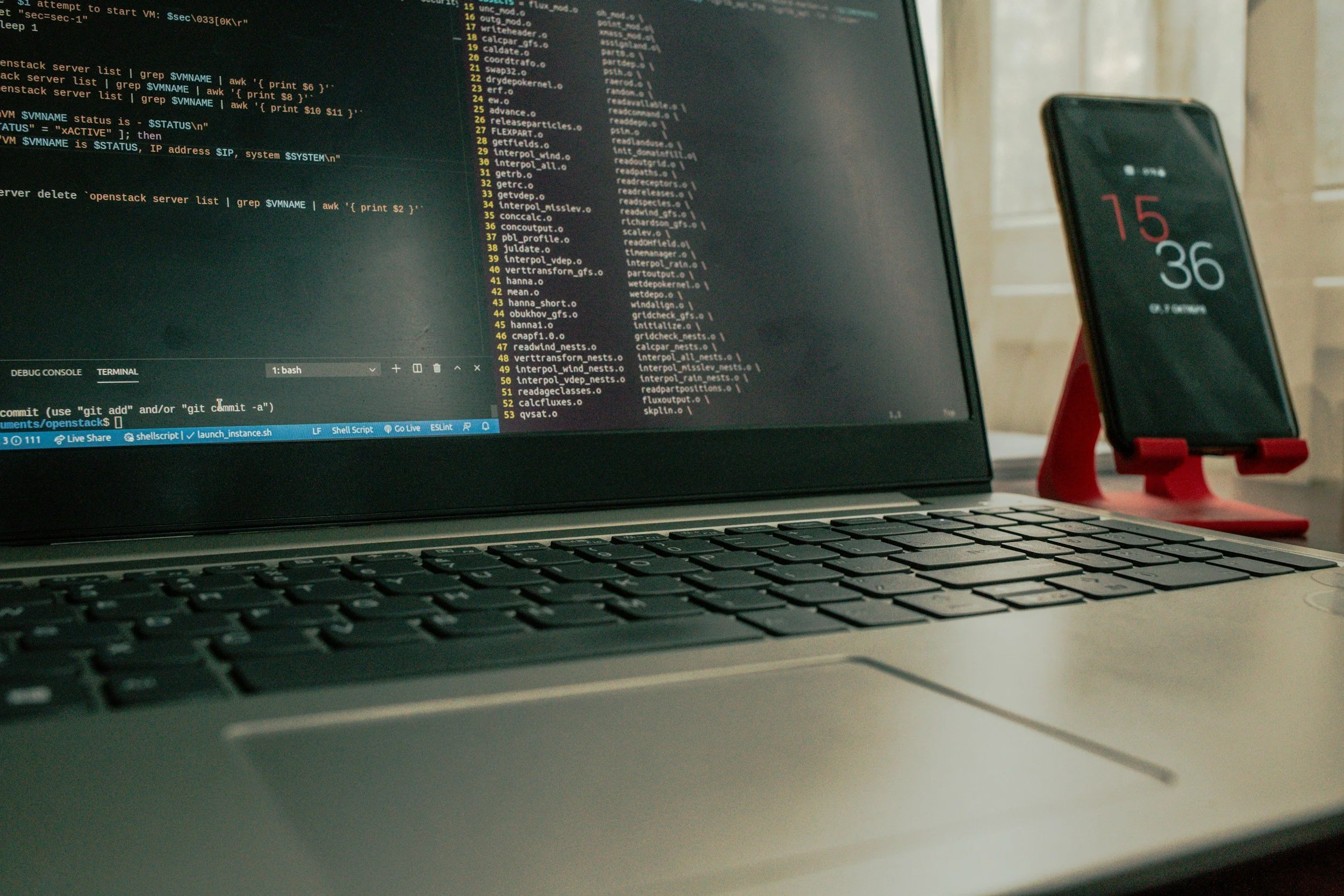 A close-up of a laptop displaying computer code, with a smartphone on a red stand in the background showing the time 15:36.