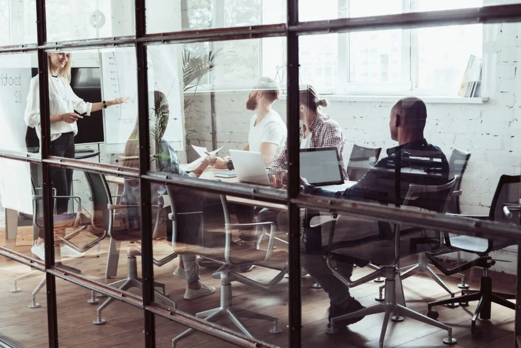A group of people in a modern office meeting through a glass wall, with one woman standing and presenting, and four others seated at a table with laptops.